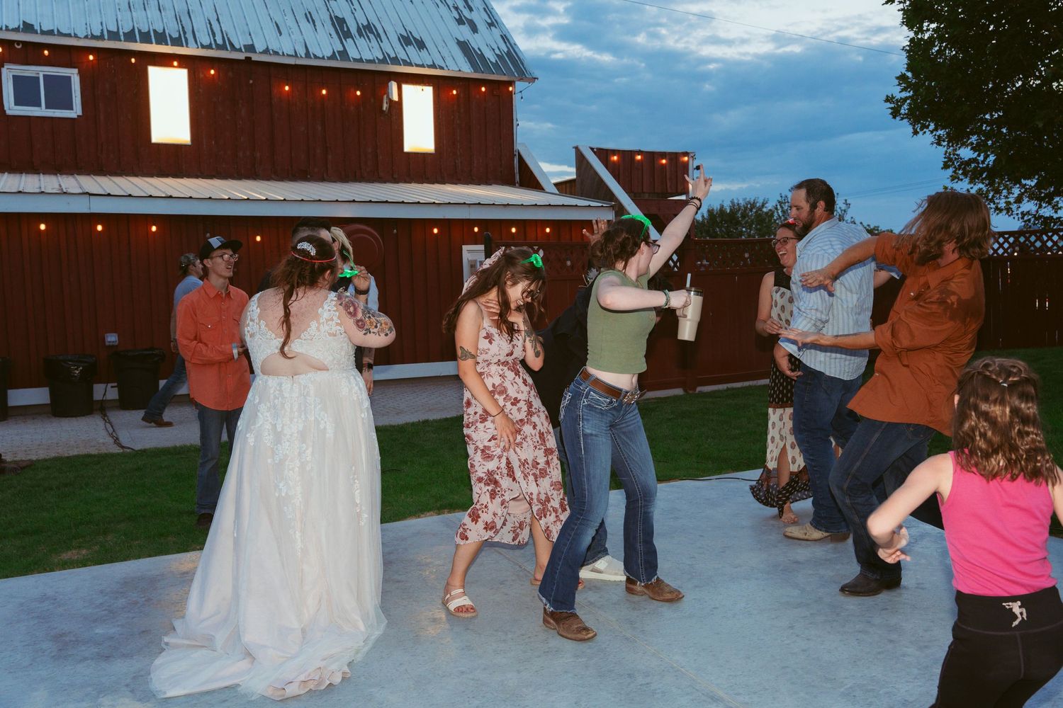 Wedding reception guests dance and socialize outside a rustic brown barn under blue skies.
