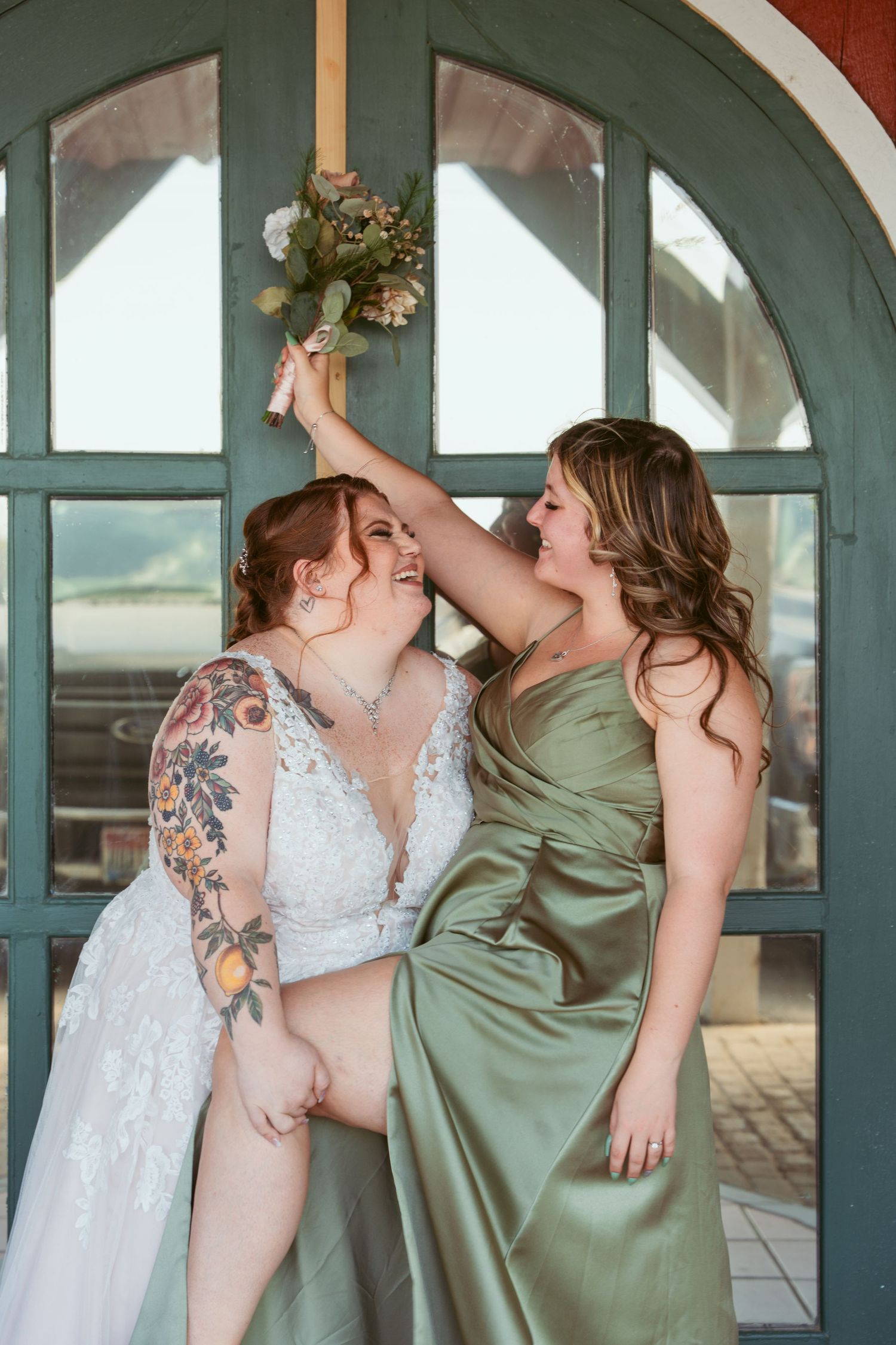 Bridesmaids in sage green dresses dance and celebrate with bride in white lace gown under arched windows.
