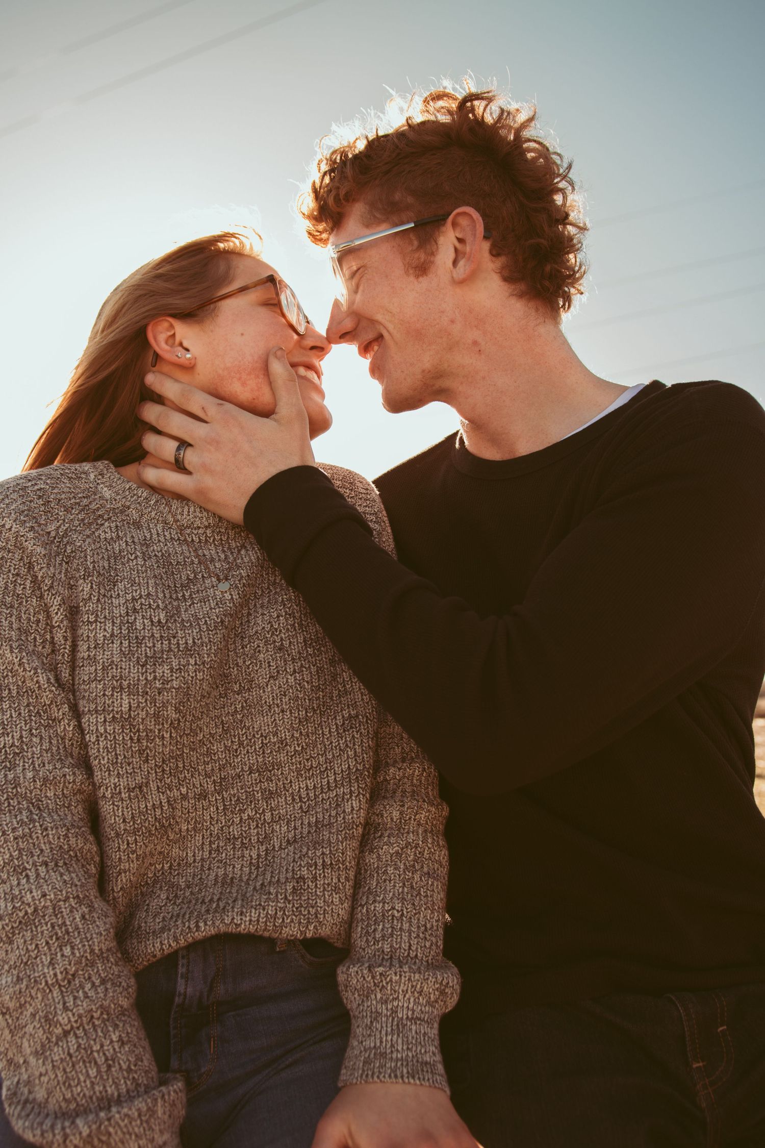 A romantic couple in casual sweaters share an intimate moment against a warm sunset backdrop.