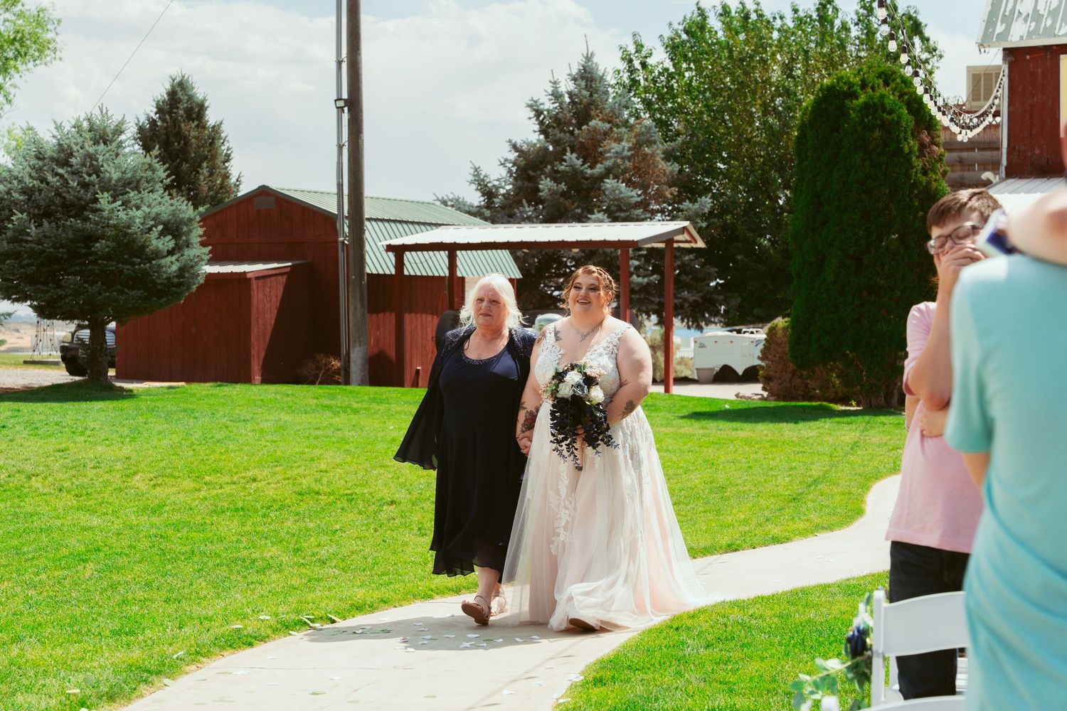Wedding ceremony procession walking down a grassy aisle with a red barn and evergreen trees in the background.