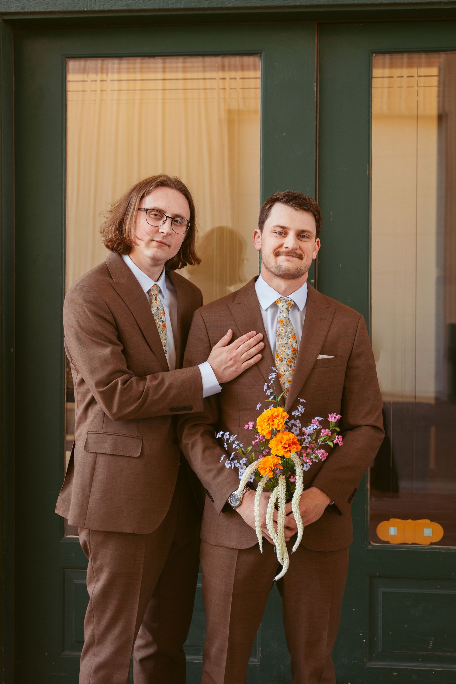 Groomsmen in brown suits pose playfully for wedding photos against brick building facade.