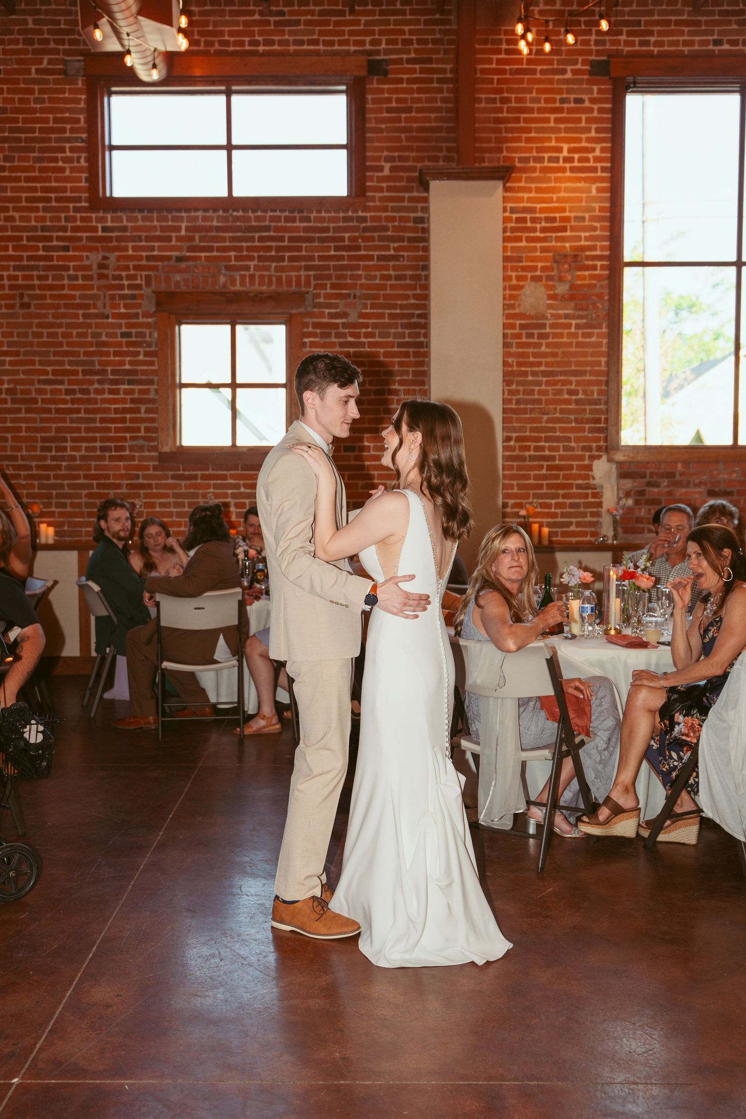 Wedding couple dances in spacious brick venue while guests watch from chairs.
