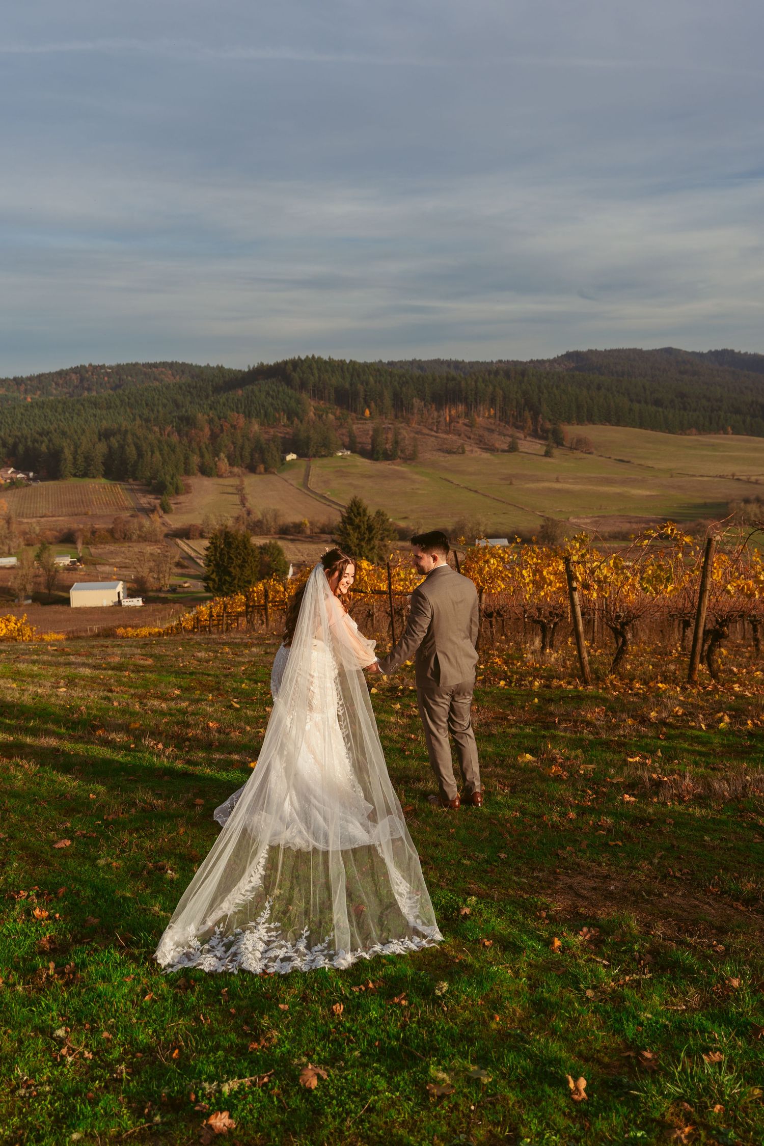 Couple walks through autumn vineyard with bride's lace veil trailing behind.