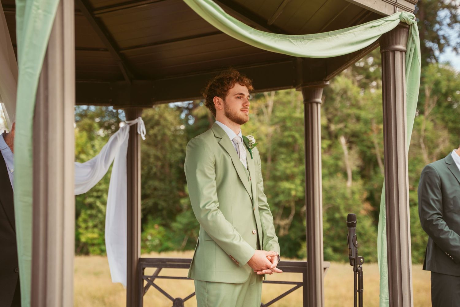 Wedding party members in mint green and pink attire stand under a rustic wooden pavilion.