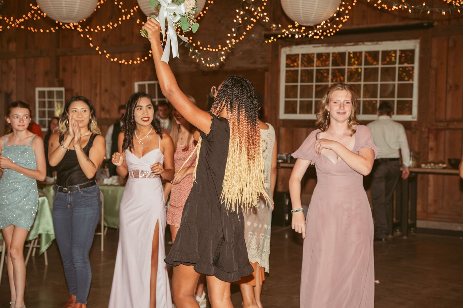 Festive wedding party dancing under decorative string lights and lanterns in a rustic barn setting.