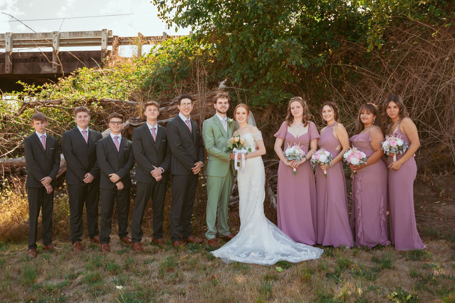 Wedding party in mauve and sage green attire poses together outdoors.