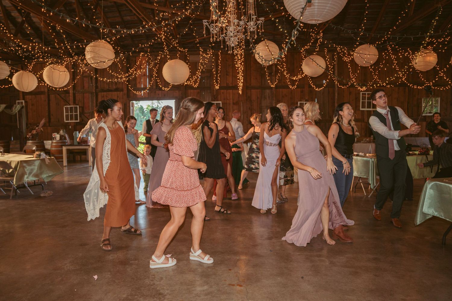Wedding guests dance under twinkling lights and paper lanterns at a rustic barn reception.