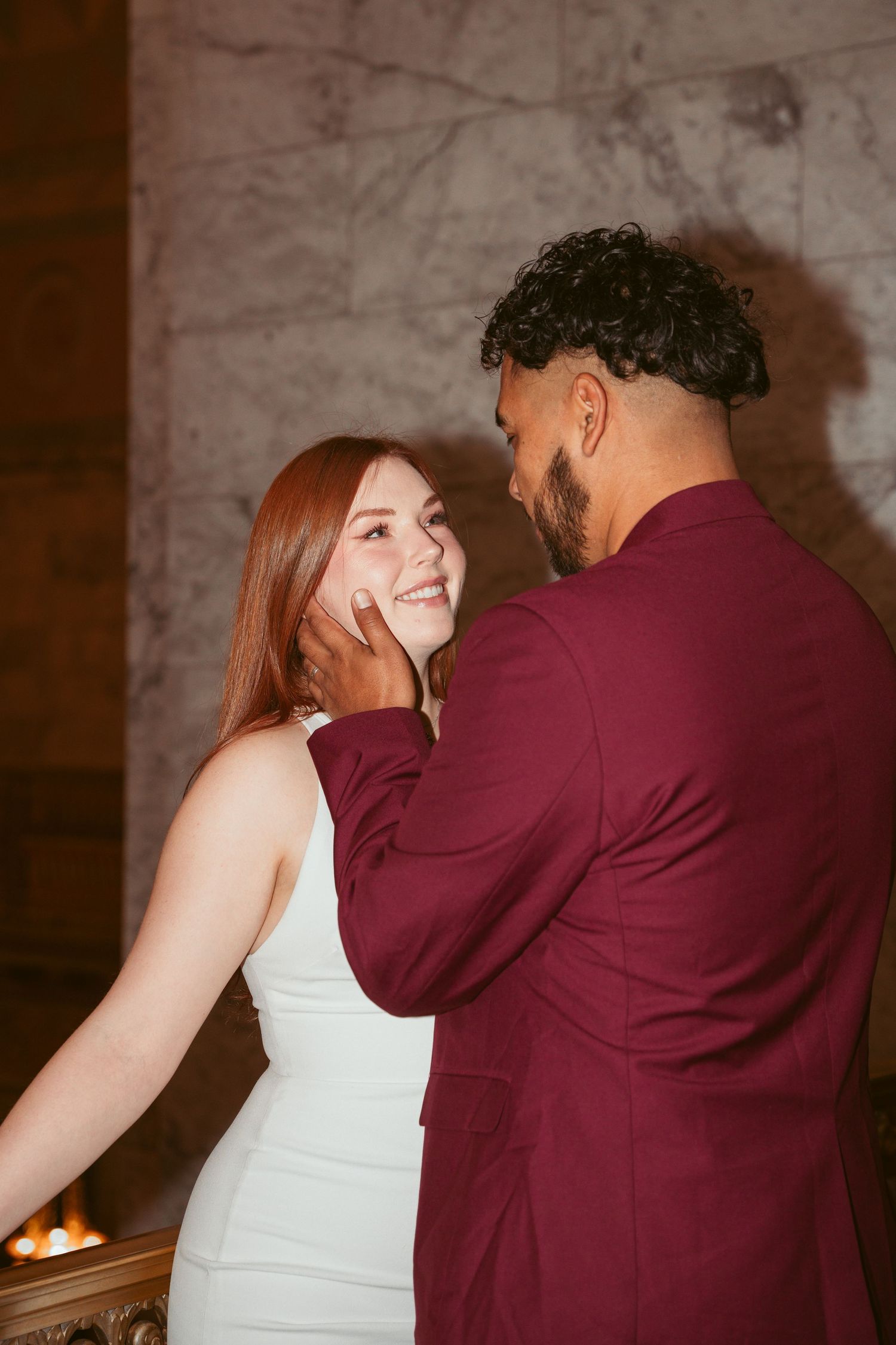 A romantic moment between a couple in burgundy and white formal attire against a marble wall backdrop.