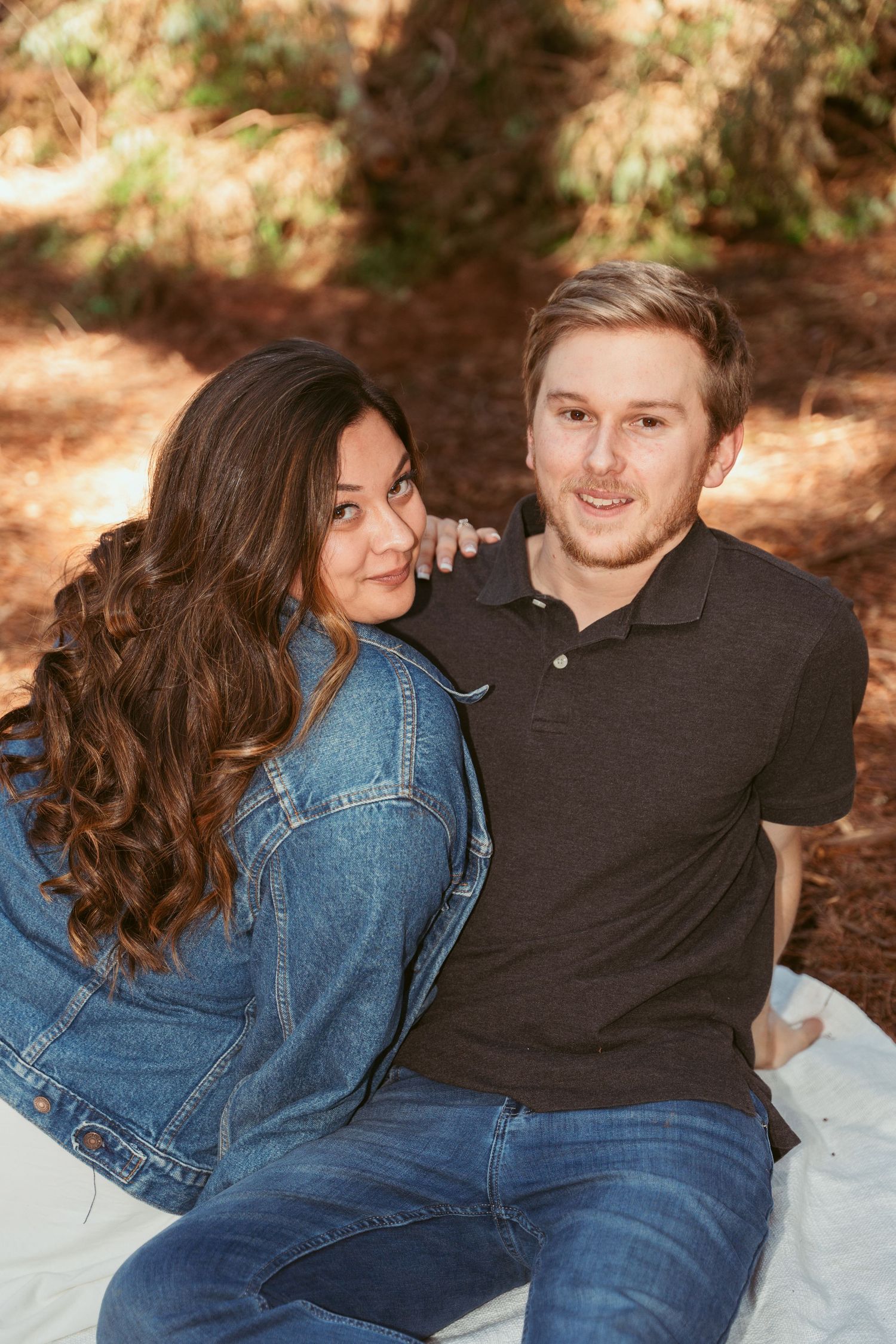 A couple relaxes on a white blanket surrounded by towering redwood trees.