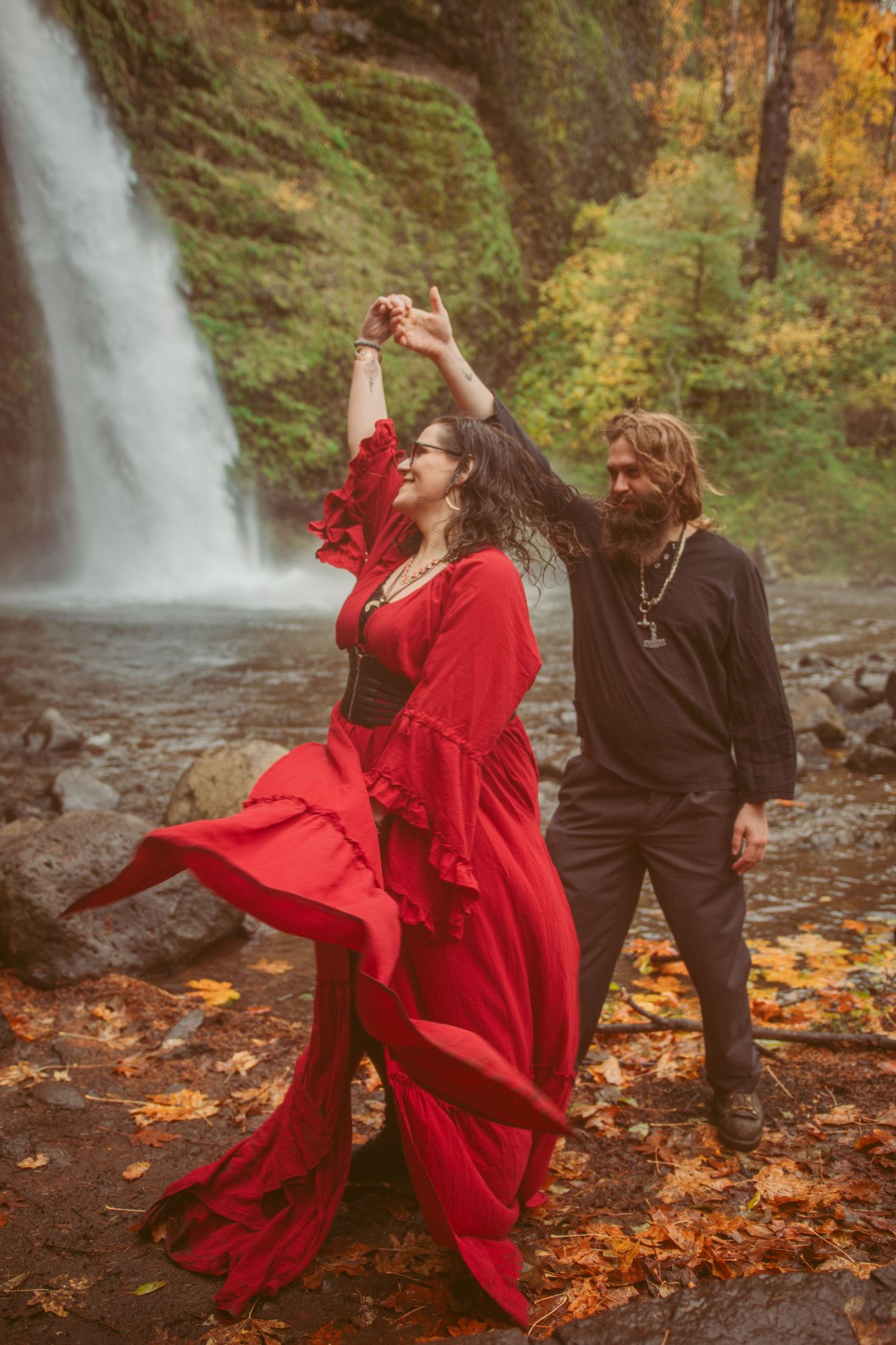A flowing red dress creates dramatic movement against autumn scenery near a cascading waterfall.