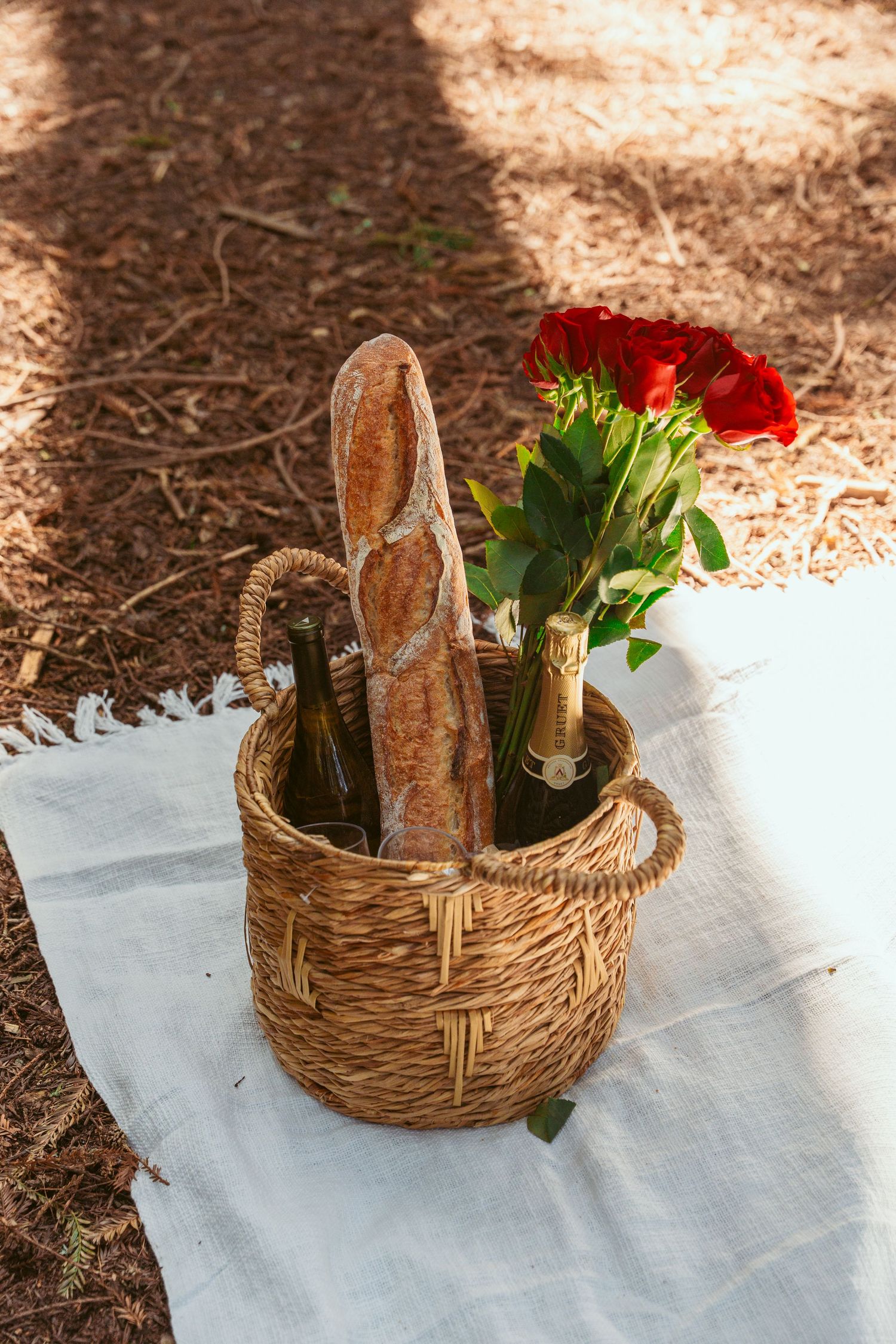 Wicker picnic basket filled with bread and flowers on a white blanket in dappled sunlight.