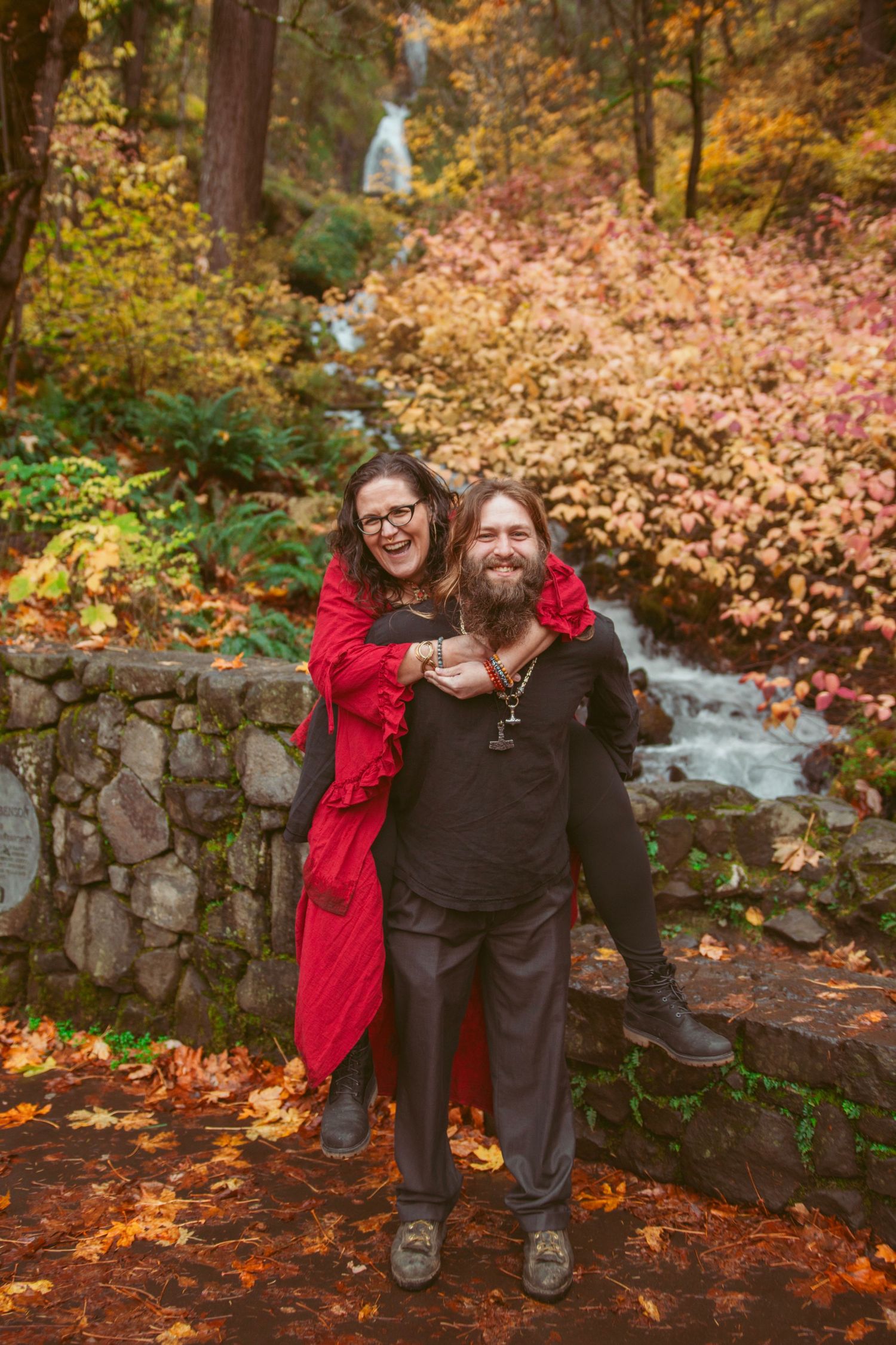 Couple shares a playful moment on fallen leaves with autumn colors and stone wall in background.