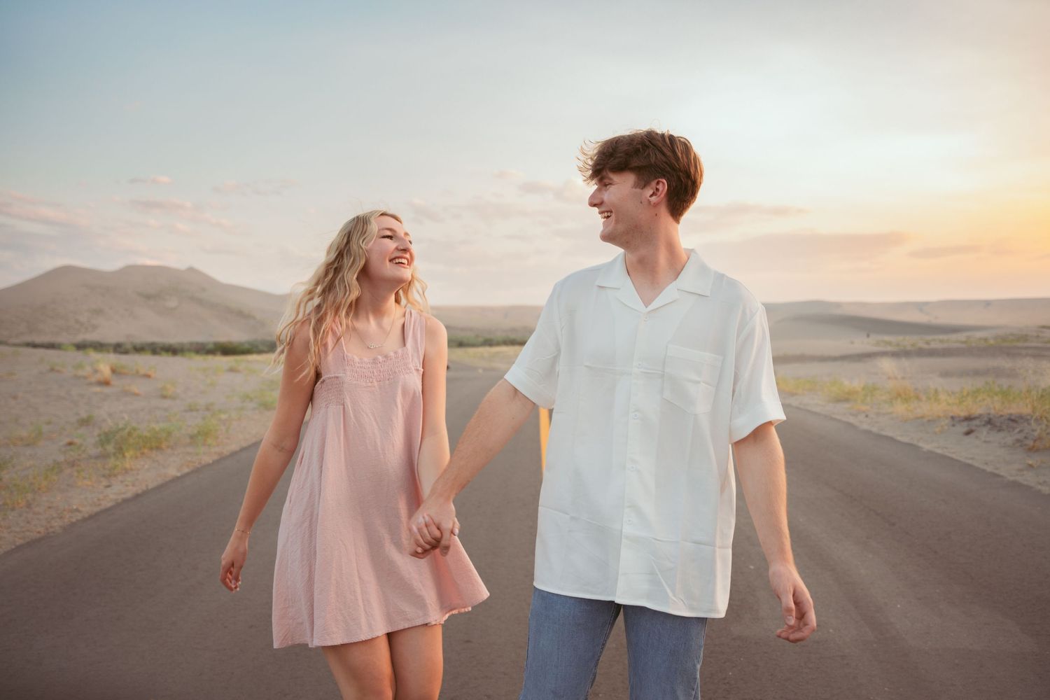 A couple in pink and white outfits walks playfully down a desert road at dusk.