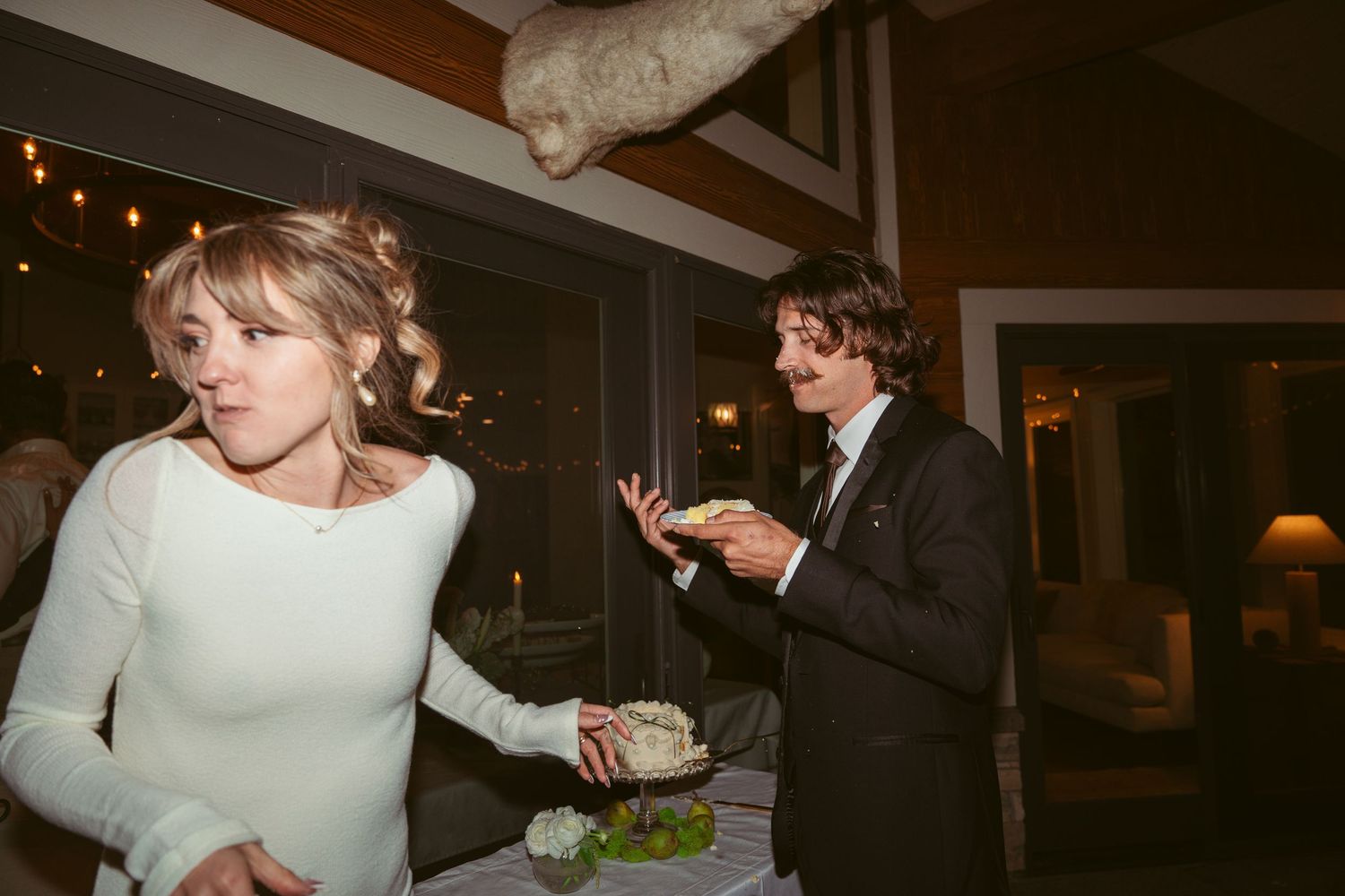A couple shares wedding cake in a dimly lit reception venue with string lights.