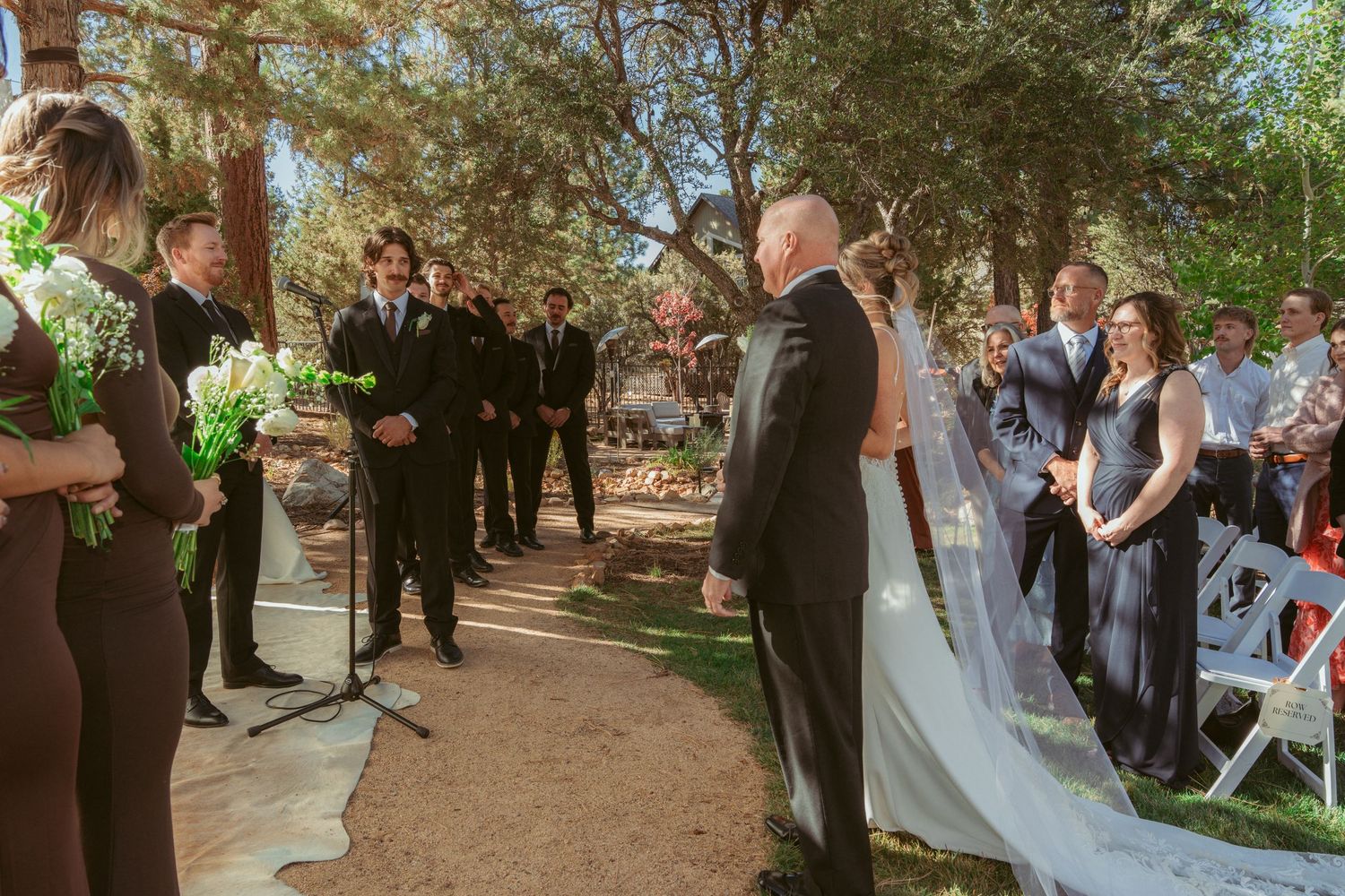 Outdoor wedding ceremony with guests gathered along a tree-lined path.