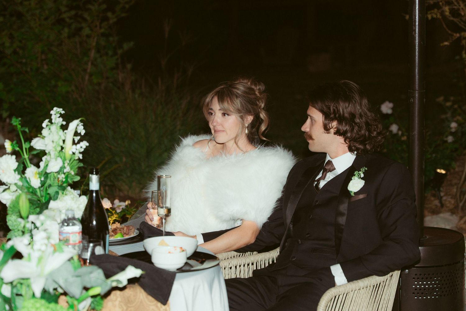 Romantic wedding reception scene with couple seated at table adorned with white floral arrangements.