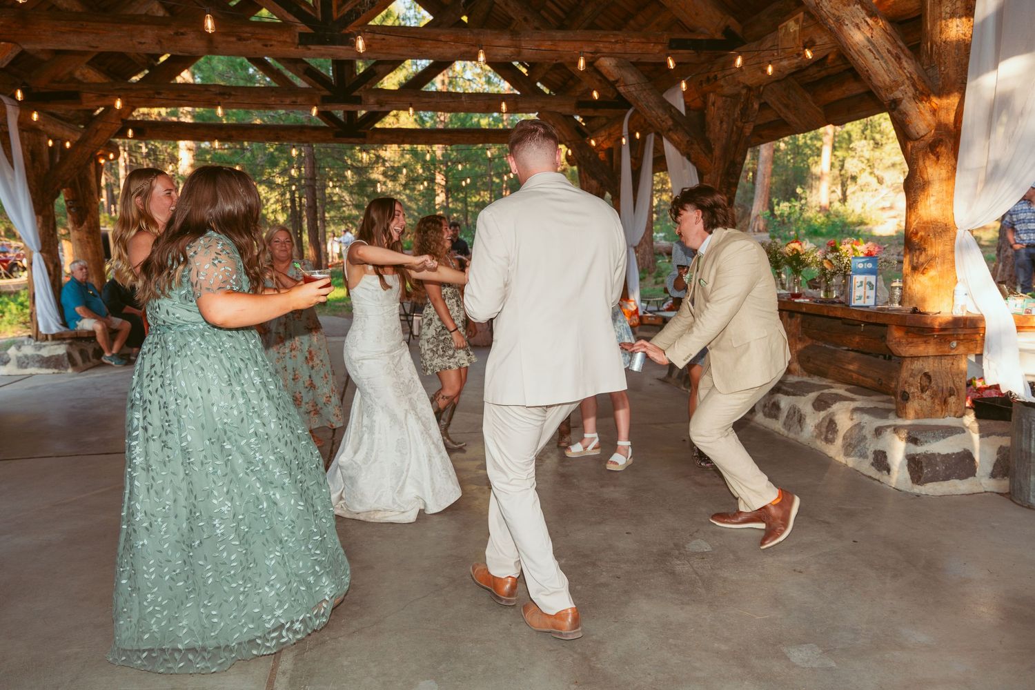 Guests enjoying line dancing at an outdoor wedding reception under wooden beams.