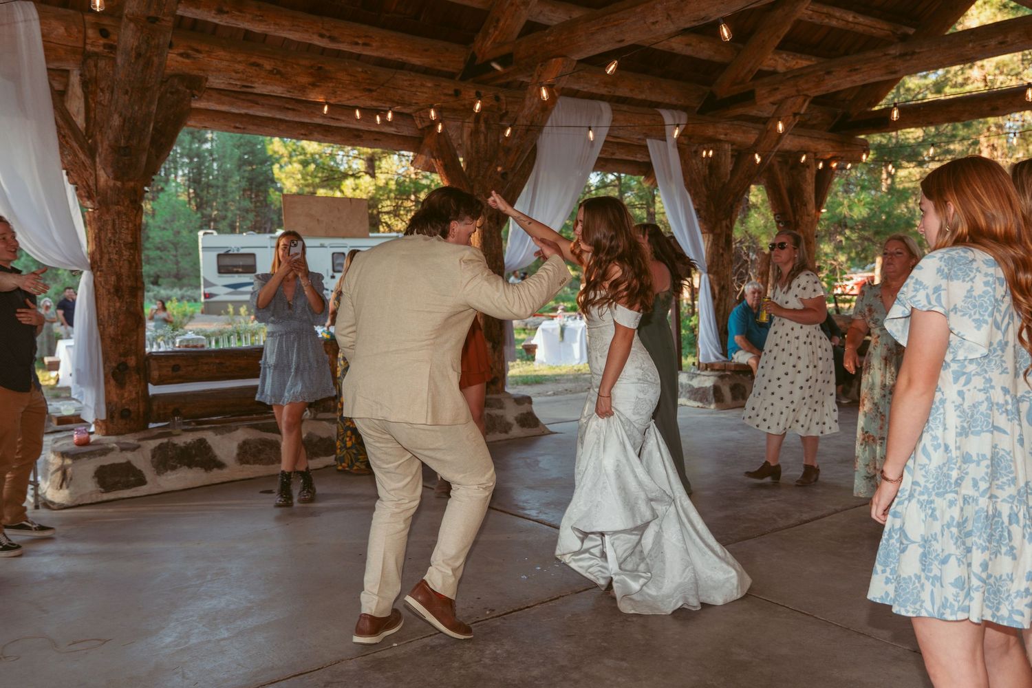 Wedding guests dance and celebrate on a rustic outdoor dance floor under string lights at sunset.