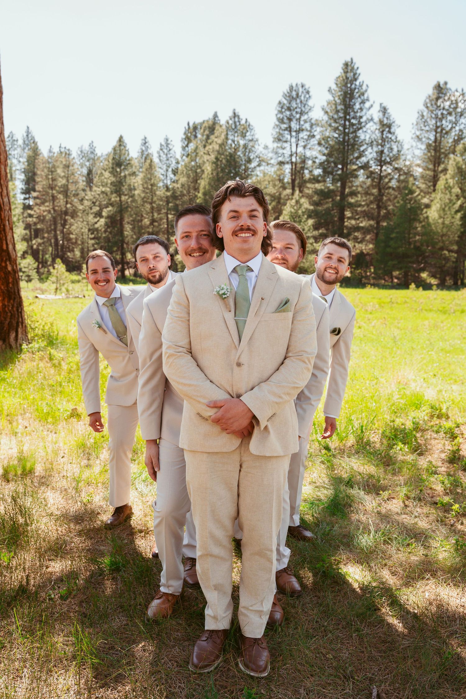 Wedding party members in beige suits share candid moments between pine trees.