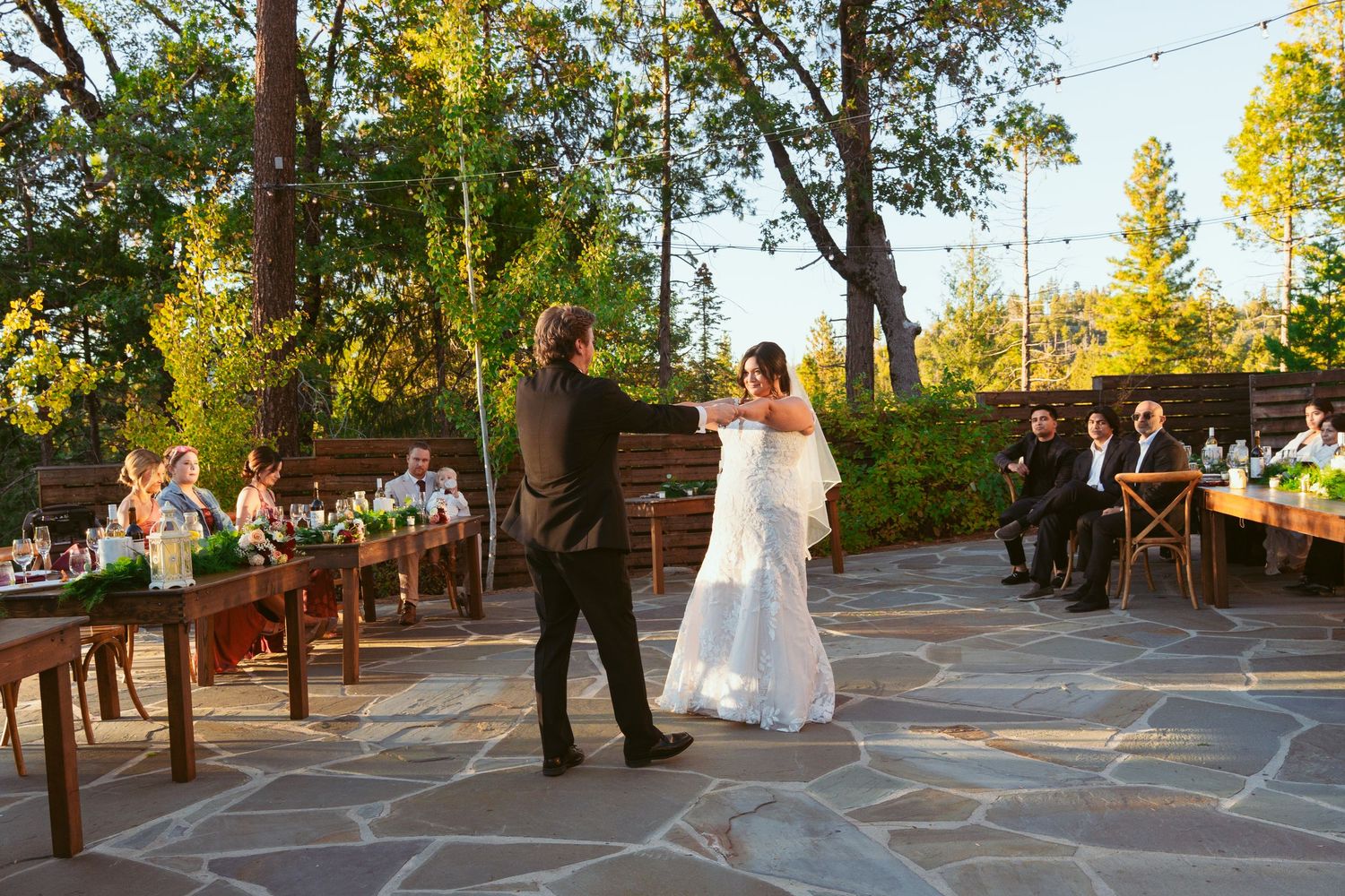 Wedding couple performs first dance on stone patio deck with wooden railings at golden hour.