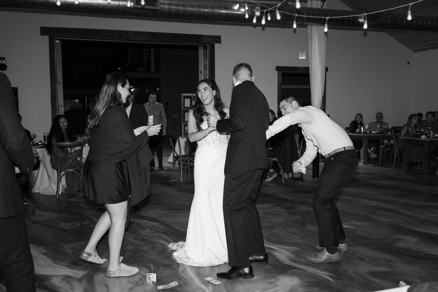 Black and white photo of wedding guests dancing at a reception with overhead lighting.