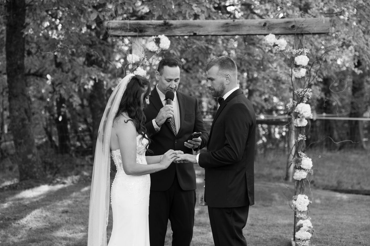 Black and white photo of wedding ceremony under decorated birch tree arch.