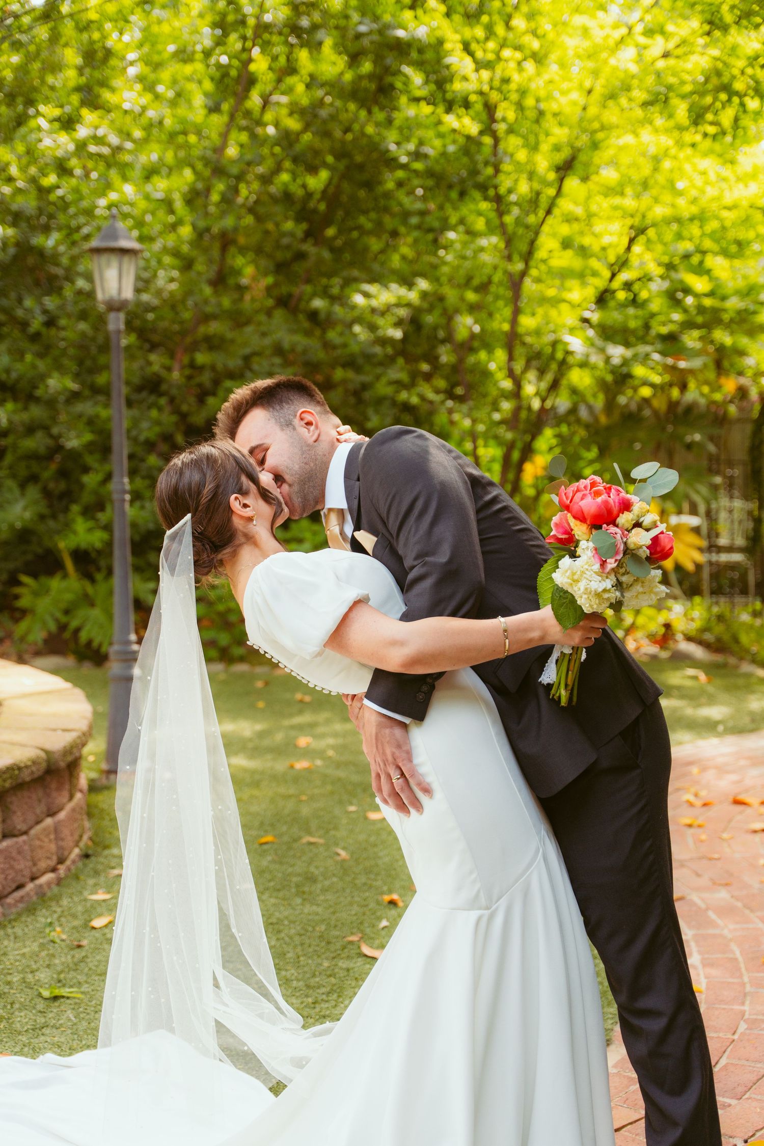 A tender wedding kiss in a sunlit garden with a flowing veil and vibrant bouquet.