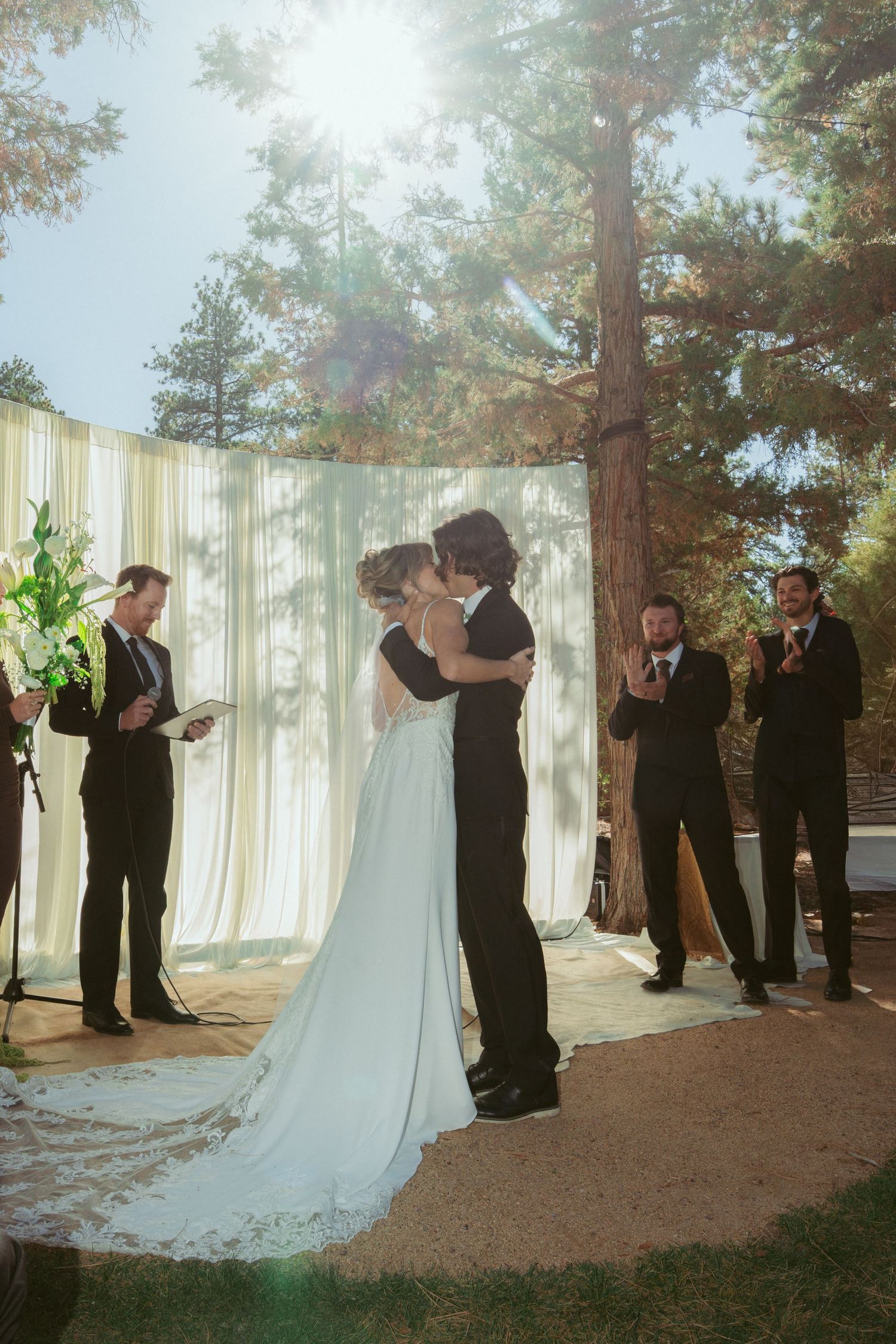 Outdoor wedding ceremony scene with white curtains and tall trees creating a natural backdrop.