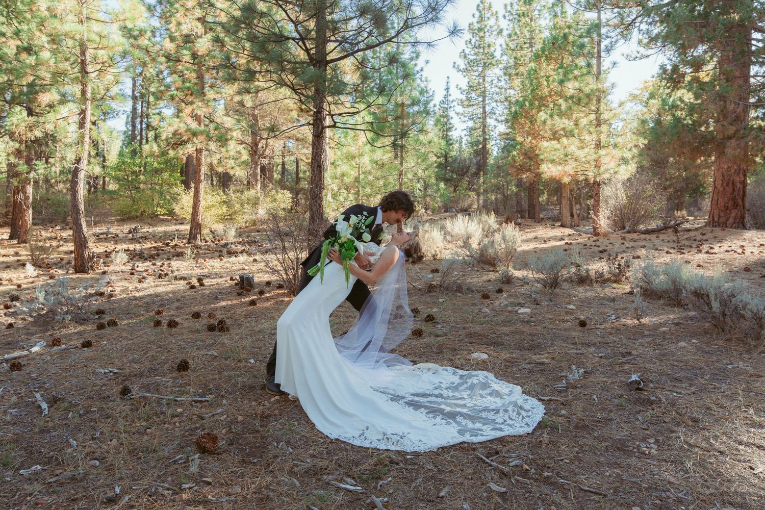 A flowing white wedding dress trails elegantly across pine needles in a forest setting.