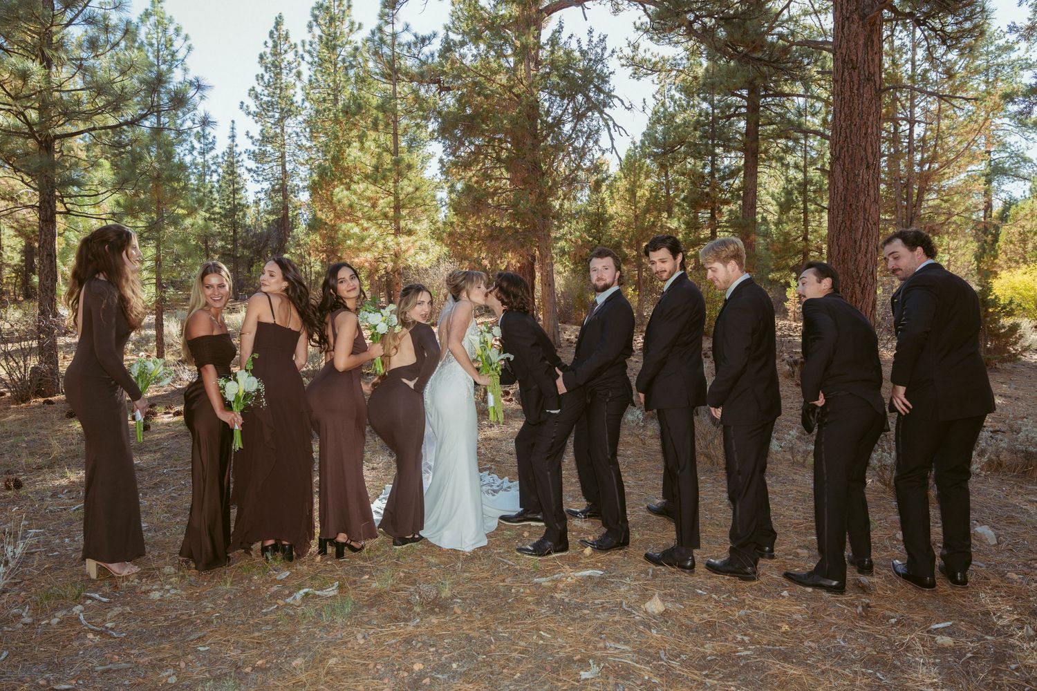 Wedding party members in brown and black attire pose together in an outdoor forest setting.
