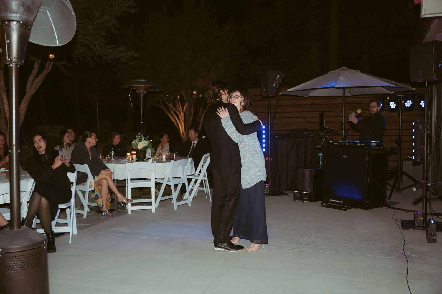 Dancers share a romantic moment on an outdoor dance floor while guests watch from white chairs.
