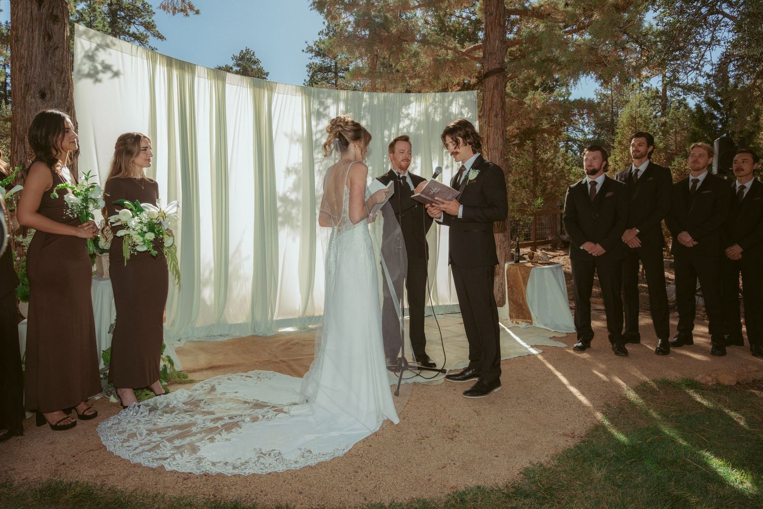 Forest wedding ceremony with white fabric backdrop and natural sunlight filtering through trees.