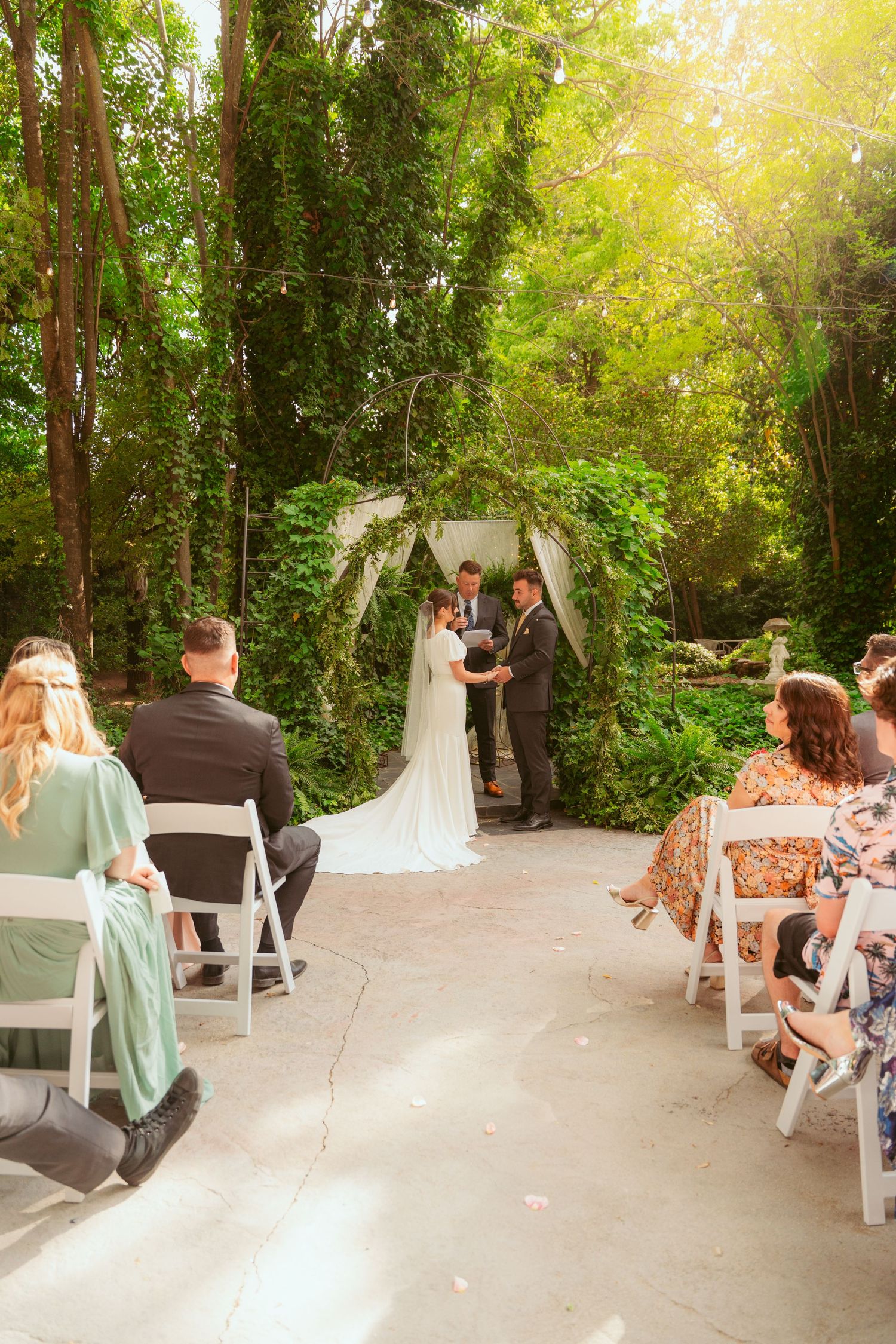 A wedding ceremony setup in a lush garden setting with white chairs and natural sunlight filtering through trees.