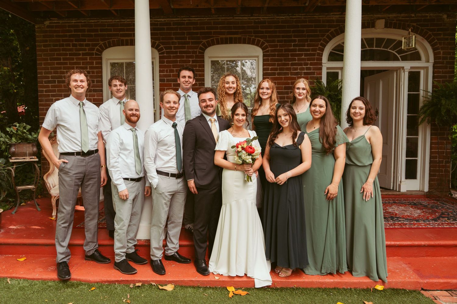 Wedding party in green dresses and gray suits gather for formal photo on red carpet.