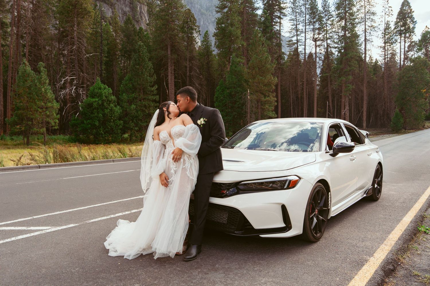 Wedding couple poses with white luxury cars on a scenic mountain road surrounded by pine trees.