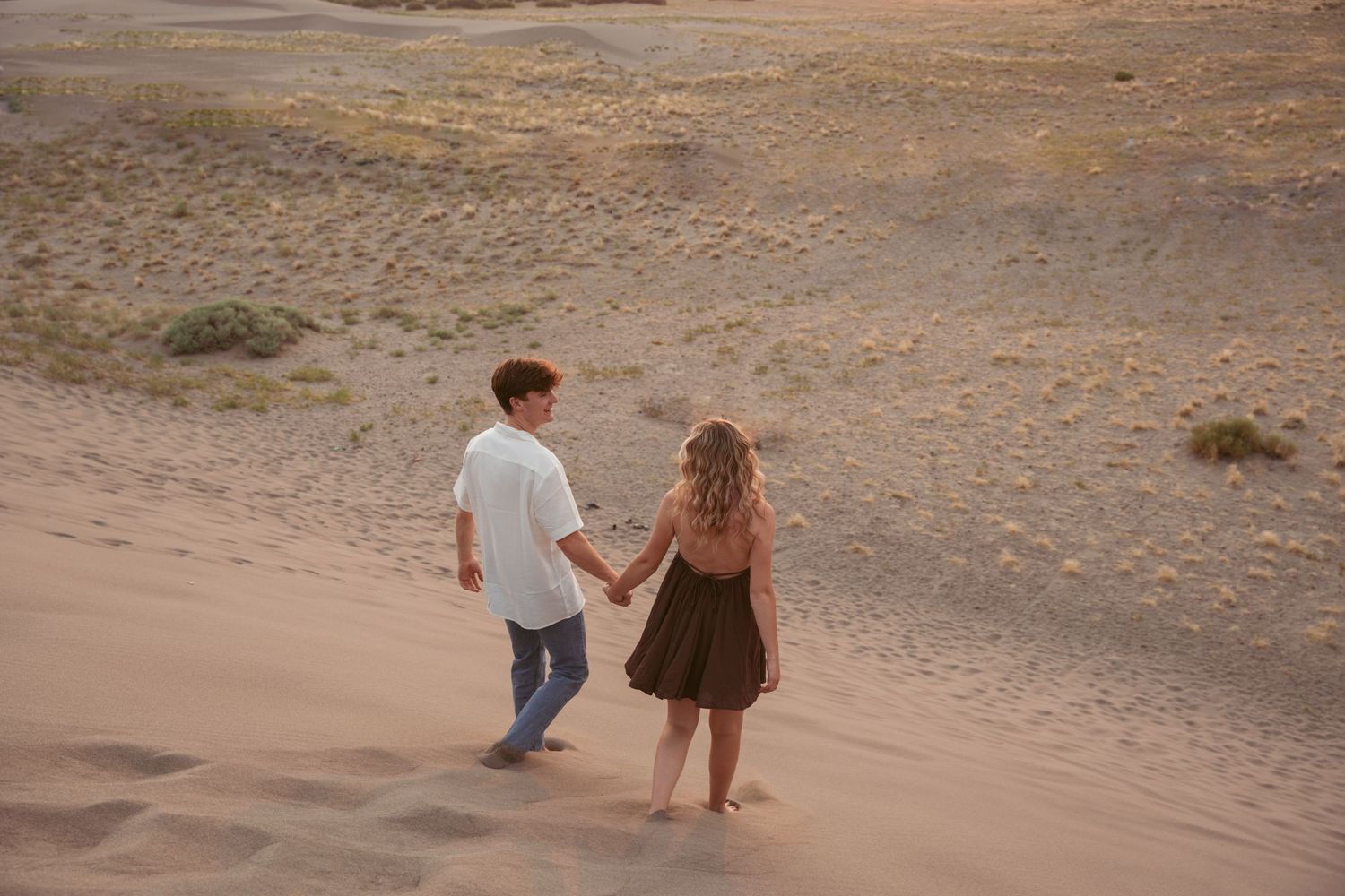 Two people walk and play together on sandy desert dunes during a sunset photoshoot.