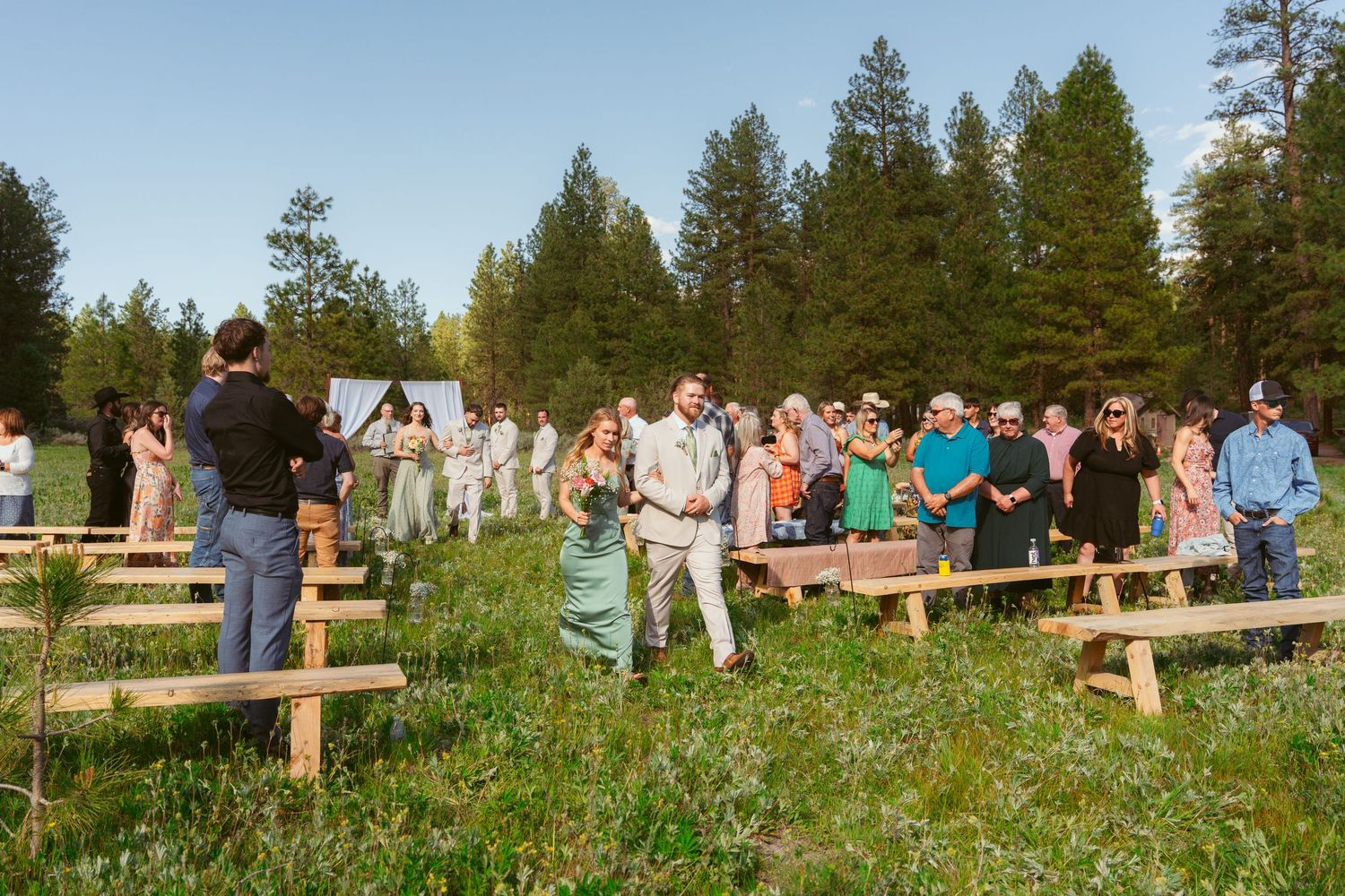 Wedding guests gather on rustic wooden benches in mountain meadow for outdoor ceremony.