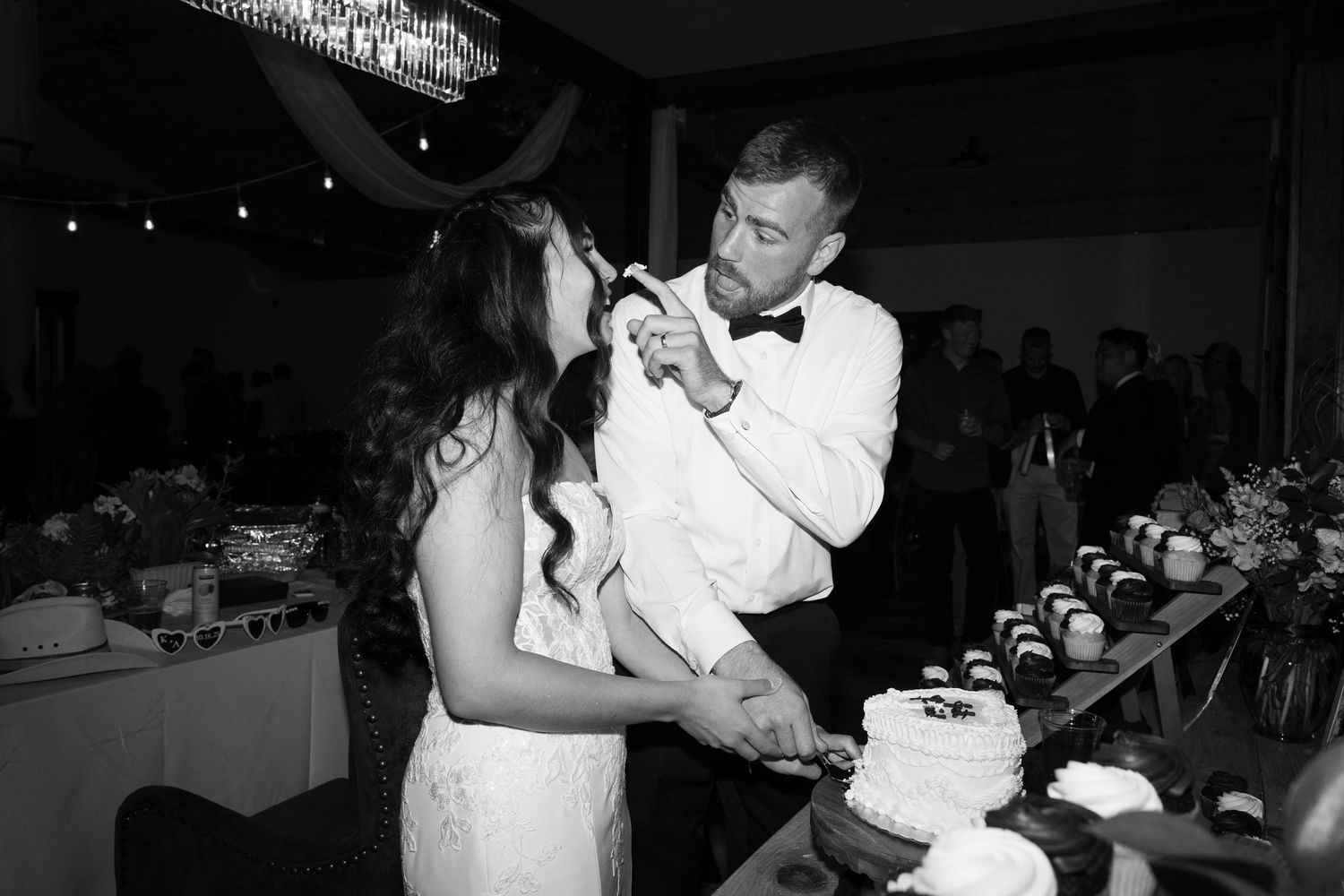 Bride and groom laugh while feeding each other wedding cake under string lights at their reception.