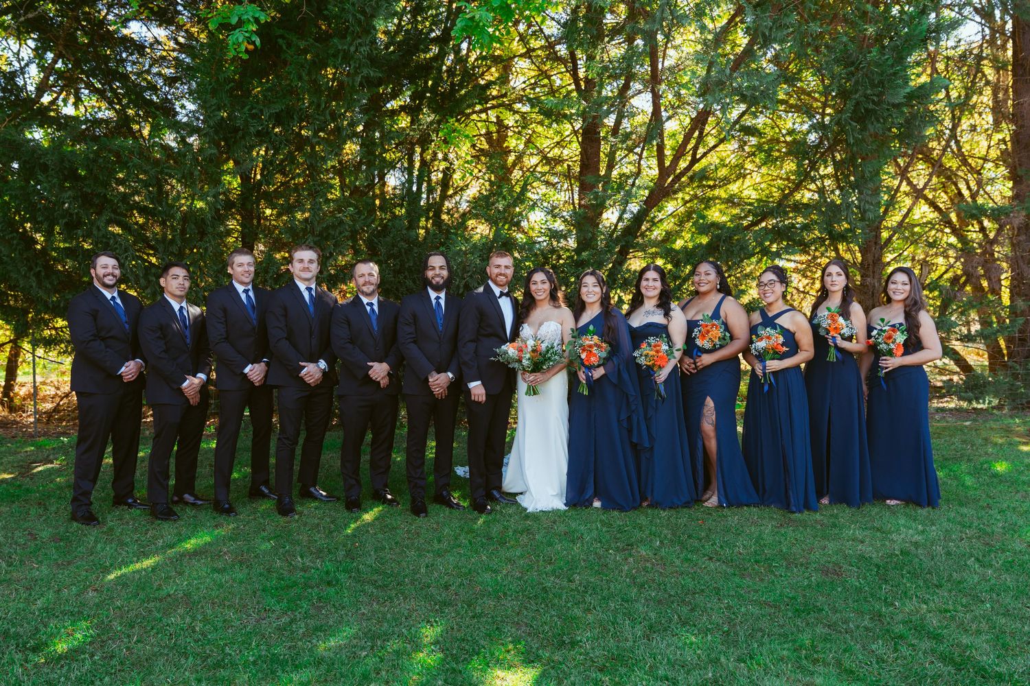 Large wedding party in navy and white attire poses together outdoors for formal group photo.