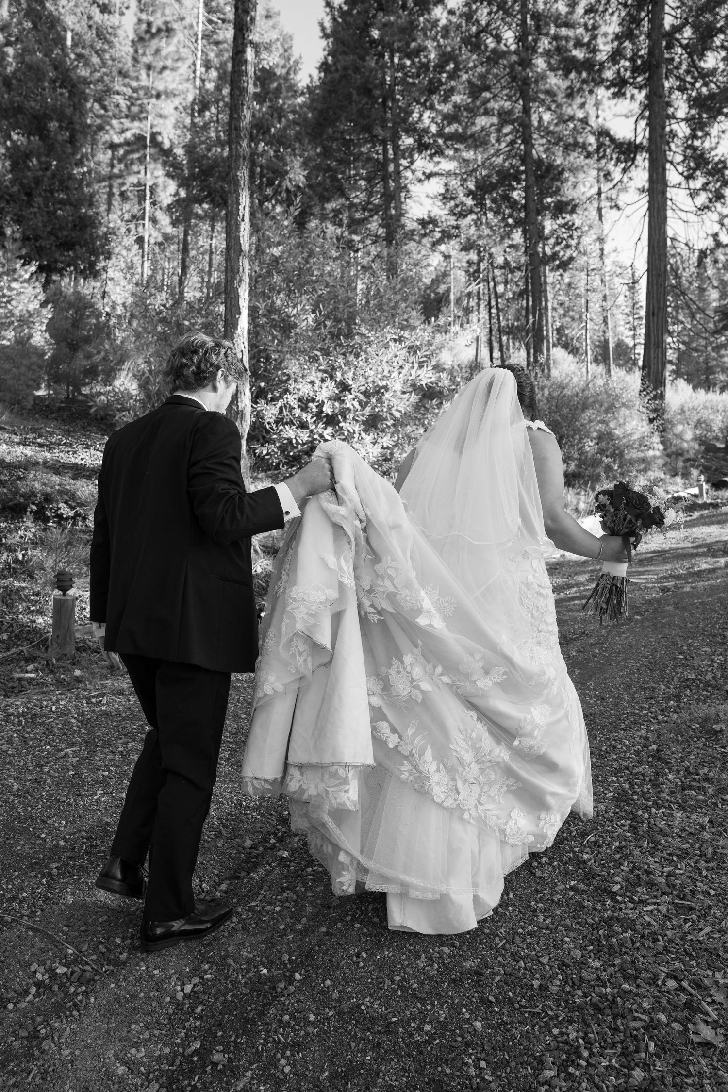 Black and white photo of couple walking along a forest path, with the groom holding the bride's train.