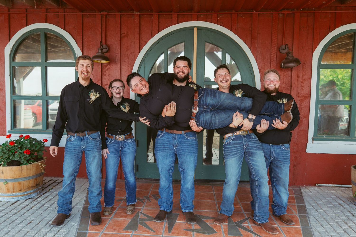 A group of men in matching dark shirts and jeans stand together on a porch of a red building.