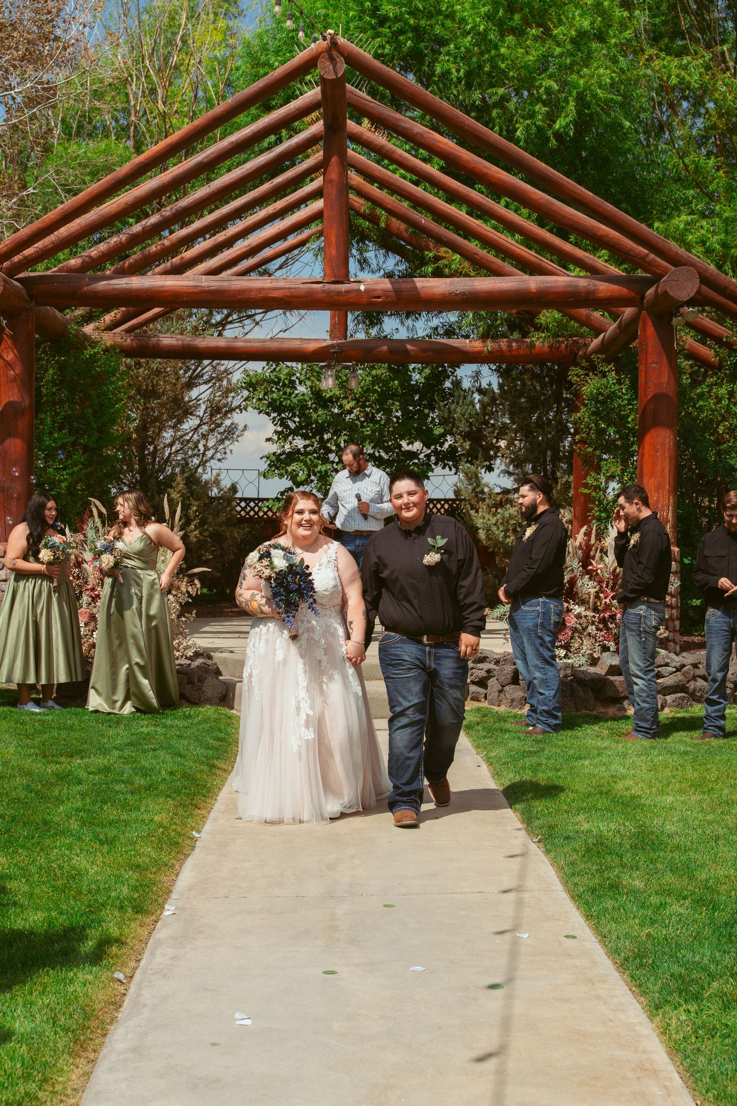 Wedding ceremony setup with wooden pergola and concrete walkway surrounded by green grass and trees.