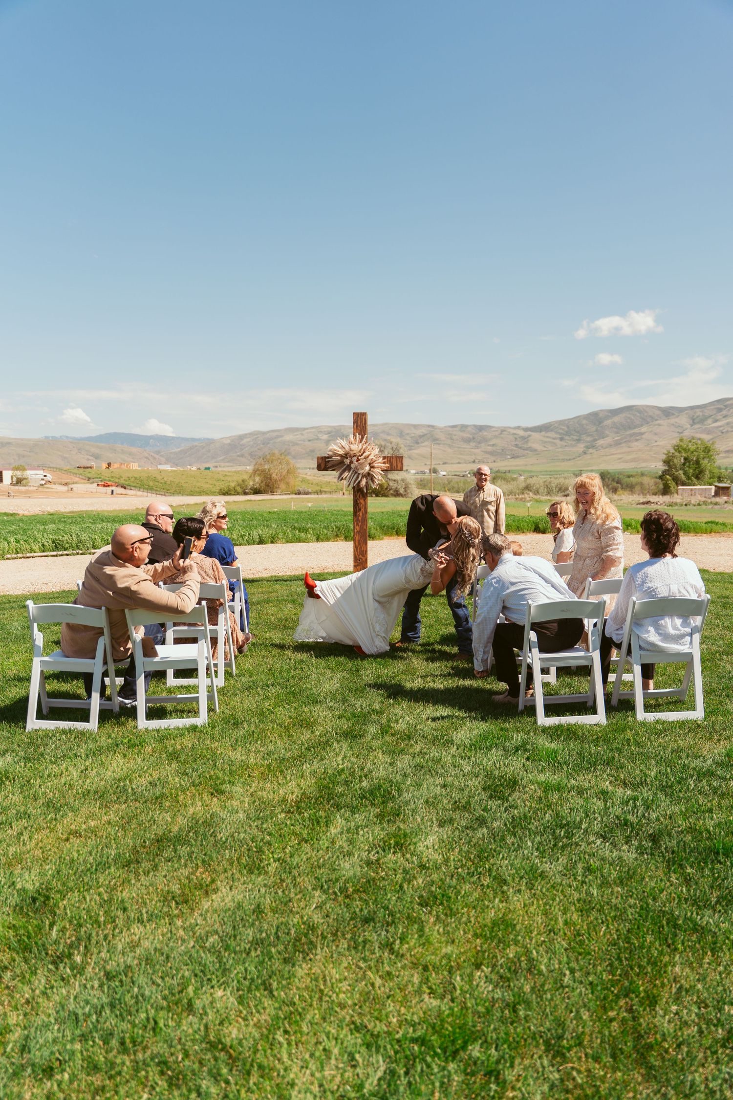Guests observe a wedding ceremony from white chairs arranged around a wooden cross on a lawn.