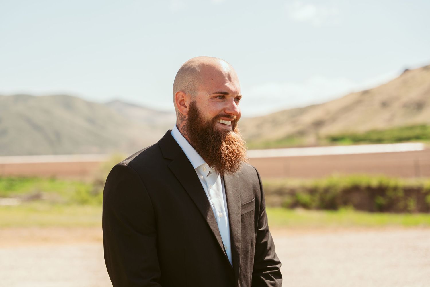 A bearded groom laughs while standing in an outdoor setting with mountains in the background.