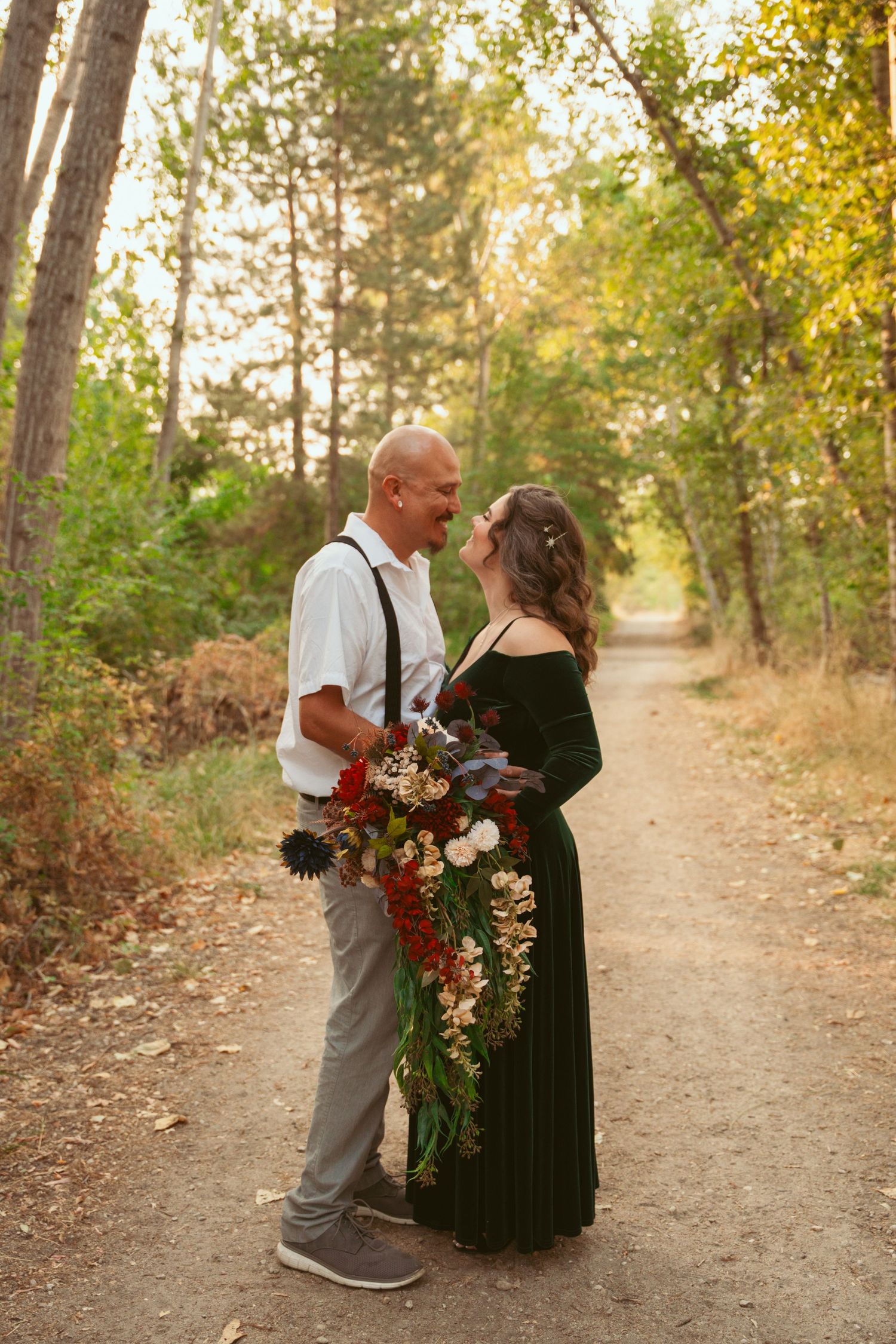 Couple in formal wear sharing an intimate moment on a forest trail with autumn flowers nearby.