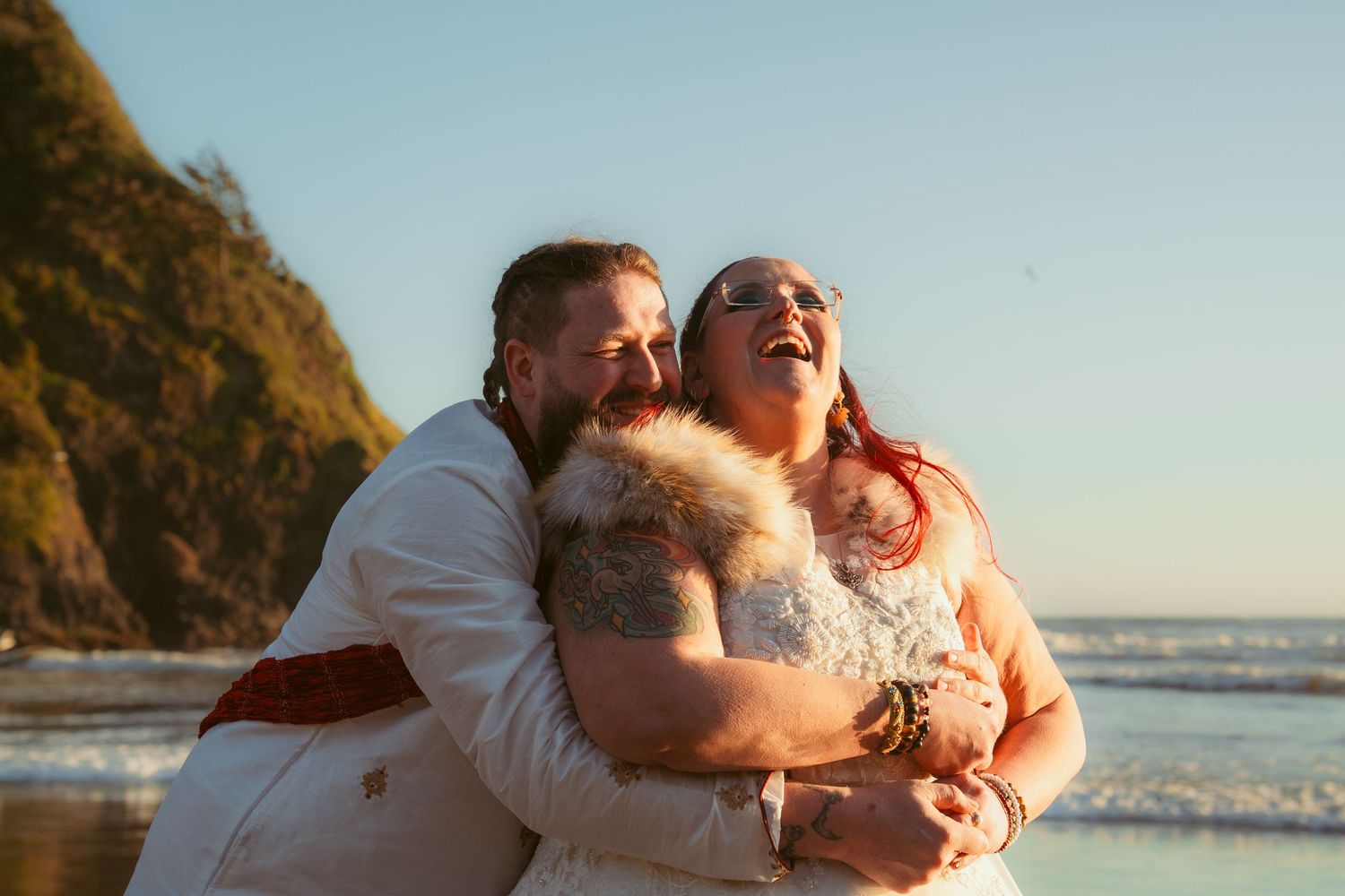 A couple in wedding attire shares an intimate moment on a beach during sunset.