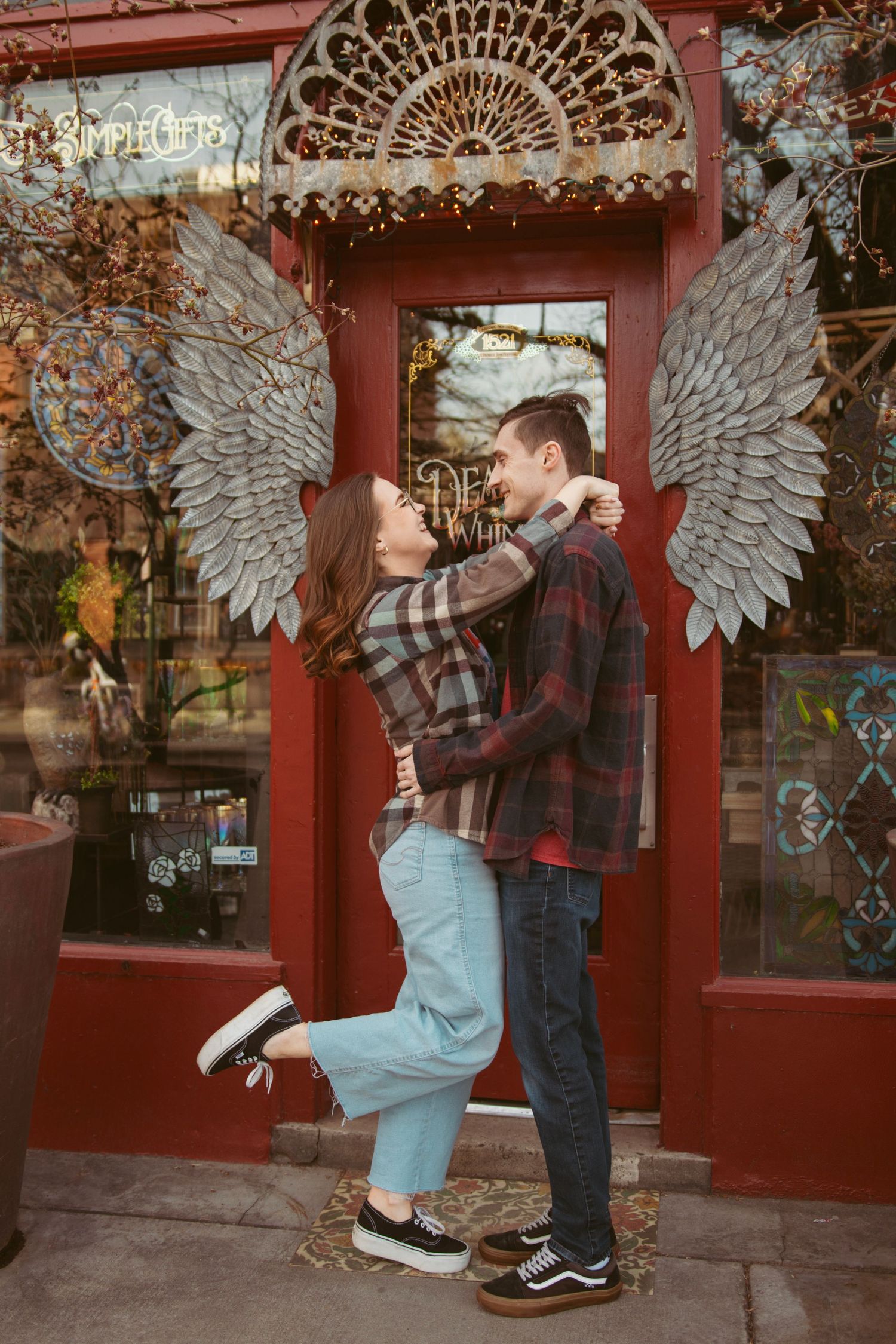 A couple poses in front of a red storefront with decorative angel wings.
