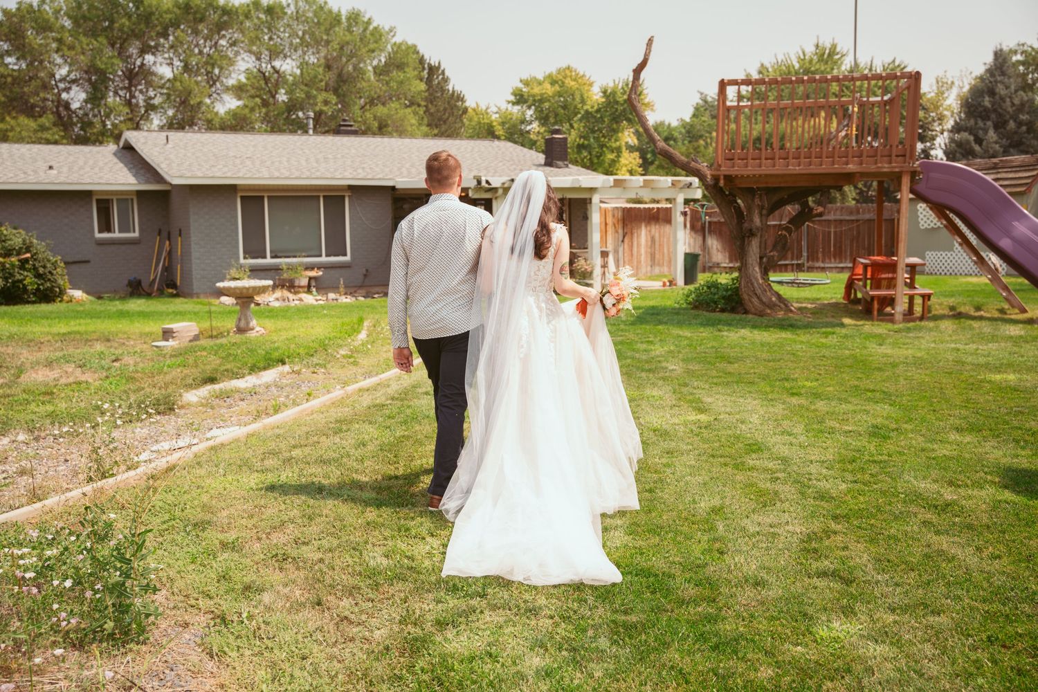 A couple walks hand in hand across a green lawn with trees and buildings in the background.