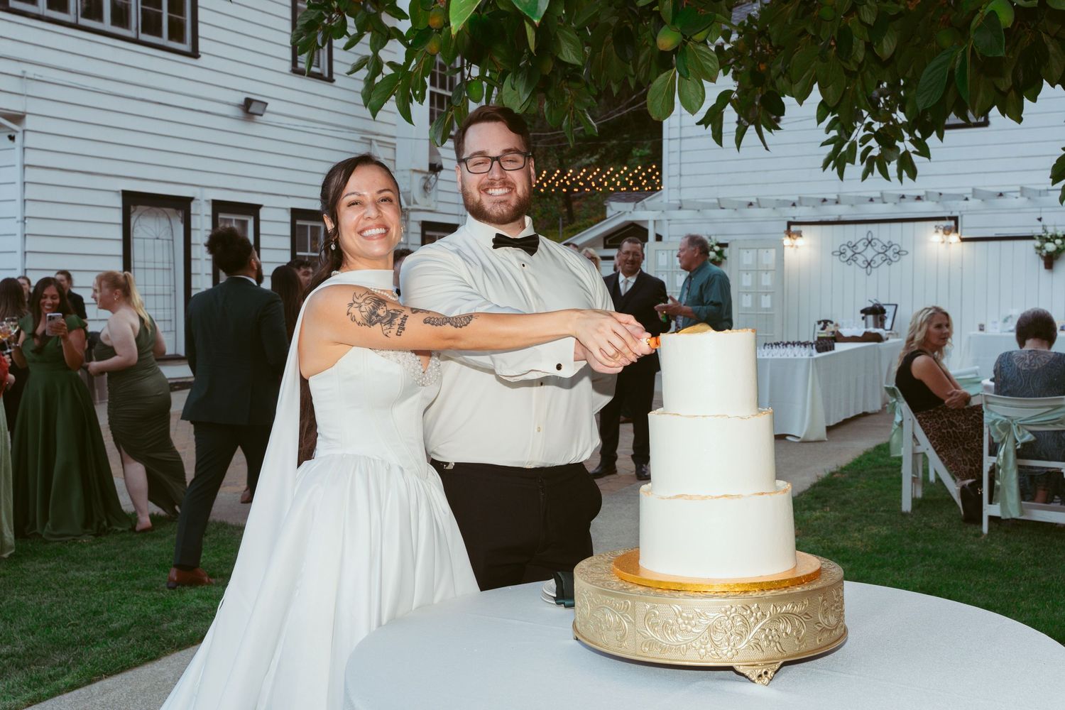 A wedding couple cuts into their elegant white and gold tiered cake at an outdoor reception.