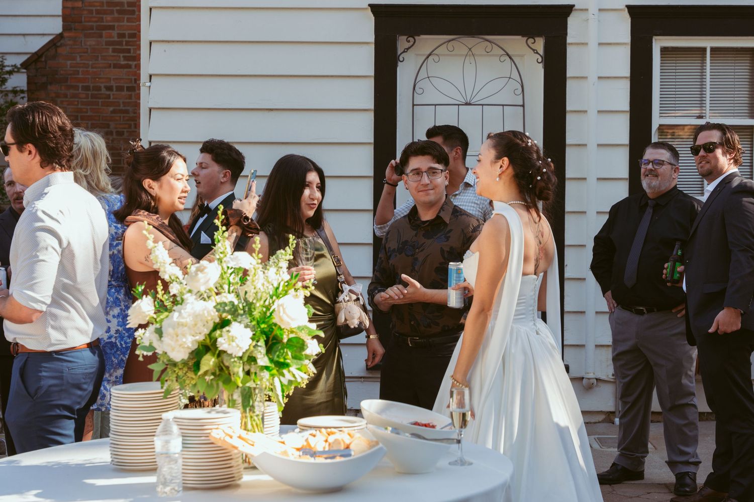 Wedding guests mingle and socialize during an outdoor reception near a white house with brick accents.